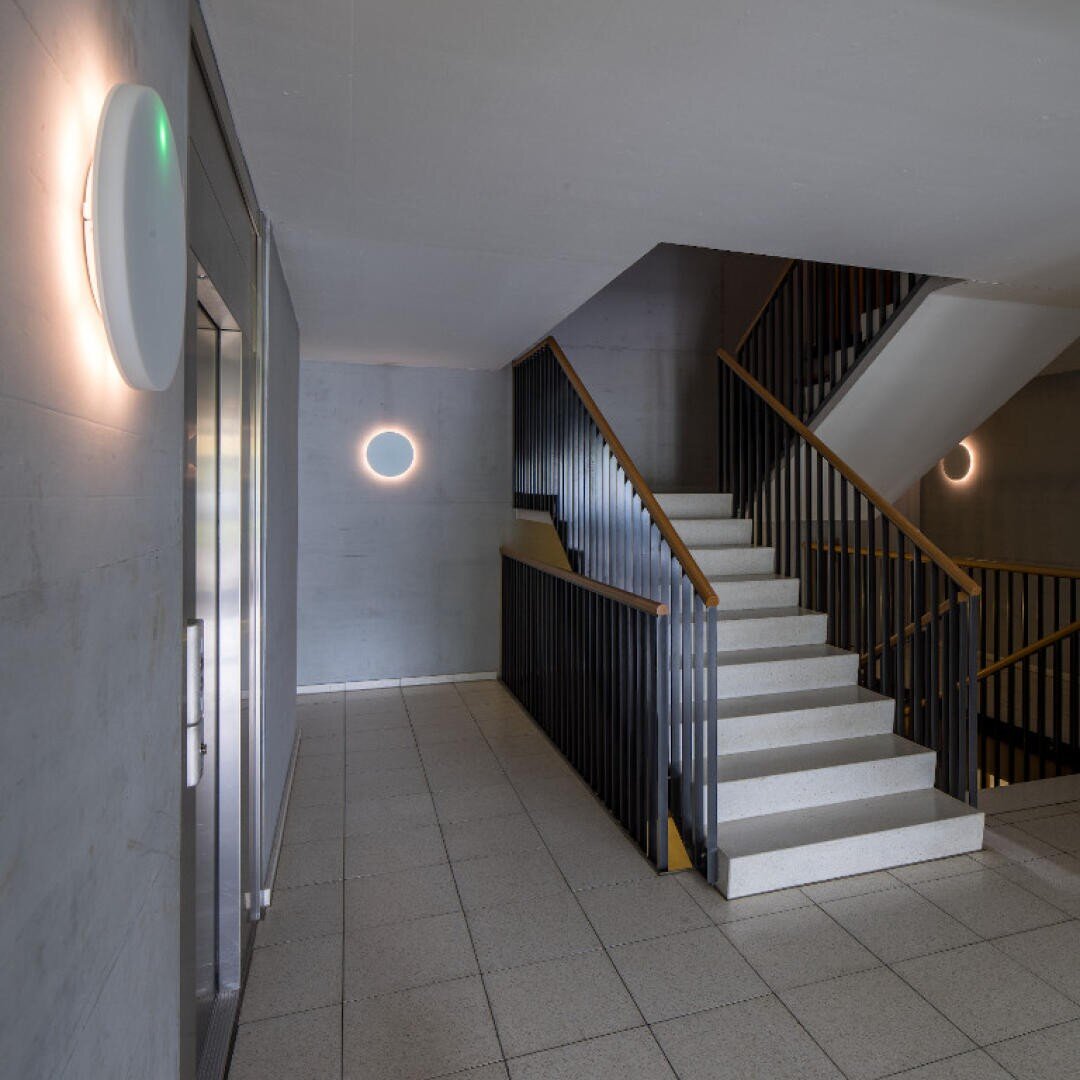 Eine moderne Innentreppe mit schwarzen Geländern und beigen Stufen, direkt neben einem Aufzug. Runde Wandleuchten geben im Grundlicht Orientierung im minimalistisch gestalteten grauen Raum, der durchgehend mit Fliesen ausgelegt ist.
