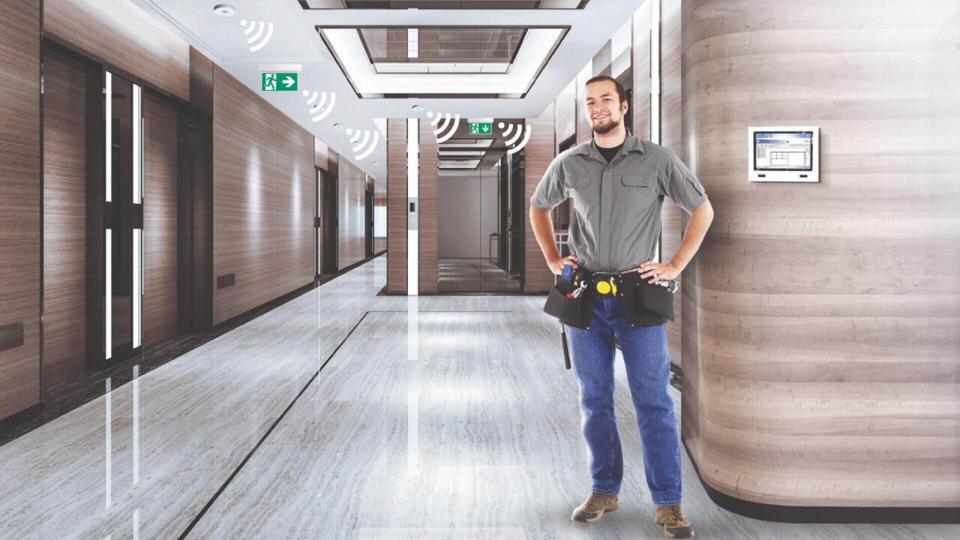 Un agent d'entretien souriant, portant une ceinture à outils, se tient dans un couloir moderne et bien éclairé, avec des murs en bois et des panneaux de sortie de secours. Des icônes de signaux sans fil sont visibles, suggérant un environnement de bâtiment intelligent.