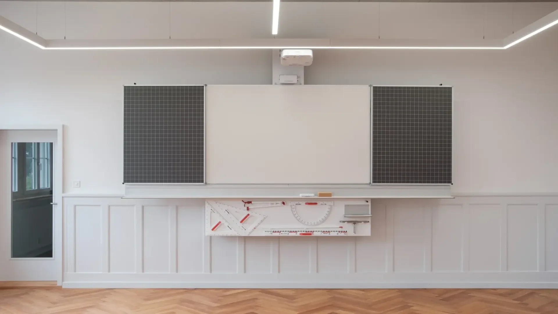 Eine Klassenzimmer-Tafel mit zwei großen schwarzen Gittertafeln an jeder Seite, einem Projektor darüber, geometrischen Hilfsmitteln darunter und einem Holzboden im Fischgrätenmuster.