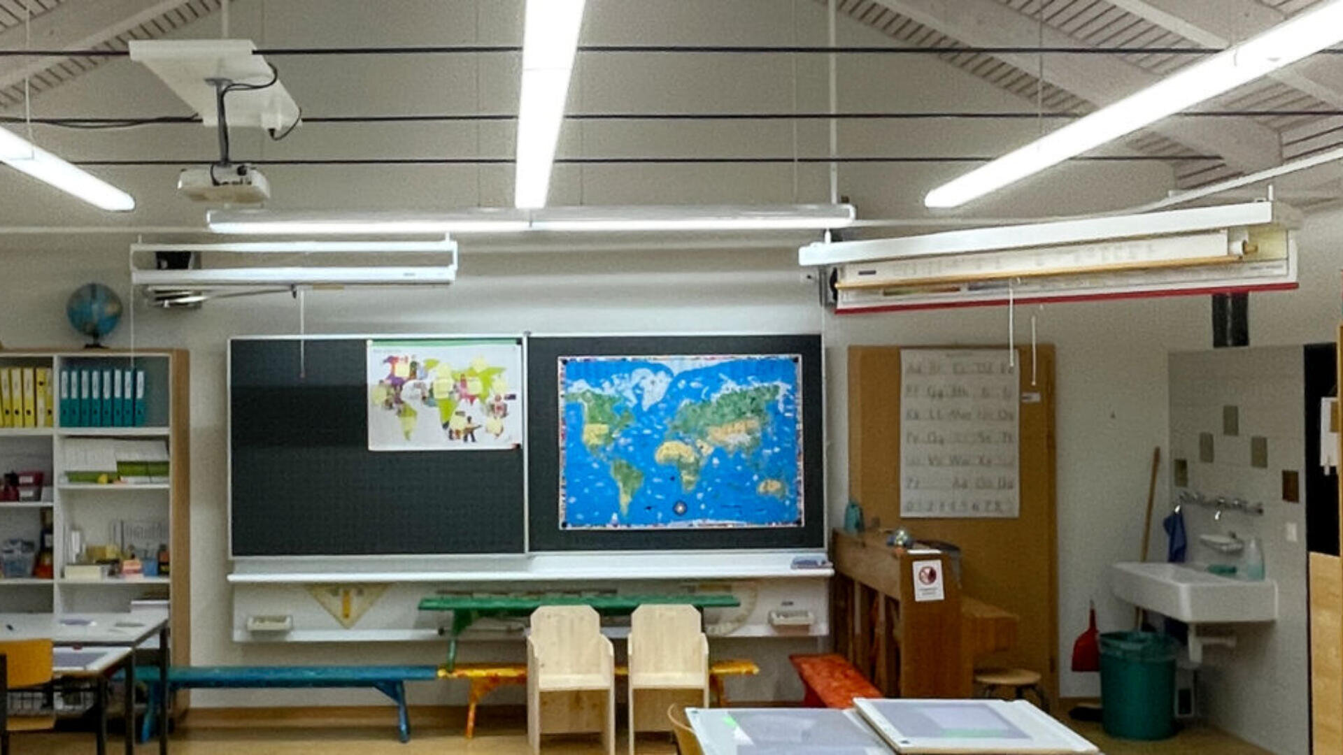 Ein hell erleuchtetes Klassenzimmer mit einer Weltkarte und Kinderzeichnungen an der Kreidetafel, Holztischen und -stühlen, bunten Regalen und einem Waschbecken in der Ecke. Die Deckenbeleuchtung und die Unterrichtsmaterialien sind sichtbar.