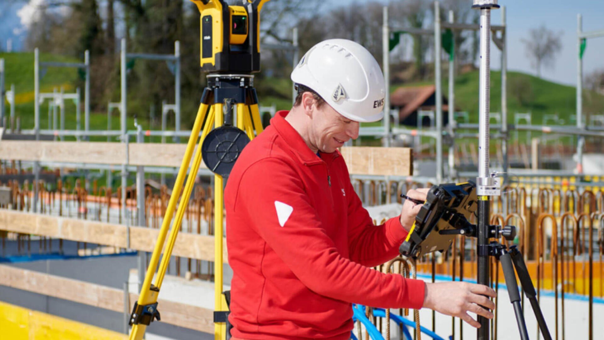 Ein Bauarbeiter in roter Jacke und weißem Schutzhelm verwendet auf einer Baustelle eine Vermessungsausrüstung und stellt ein Gerät auf einem Stativ ein. Im Hintergrund sind Metallgeländer und ein gelbes Stativ zu sehen.