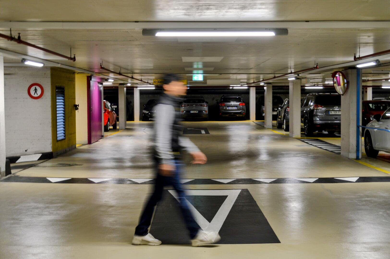 Eine sich bewegende Person geht durch eine Tiefgarage, in der auf beiden Seiten Autos parken und die fluoreszierend beleuchtet ist. Das Bild ist leicht unscharf, wodurch die Bewegung betont wird.