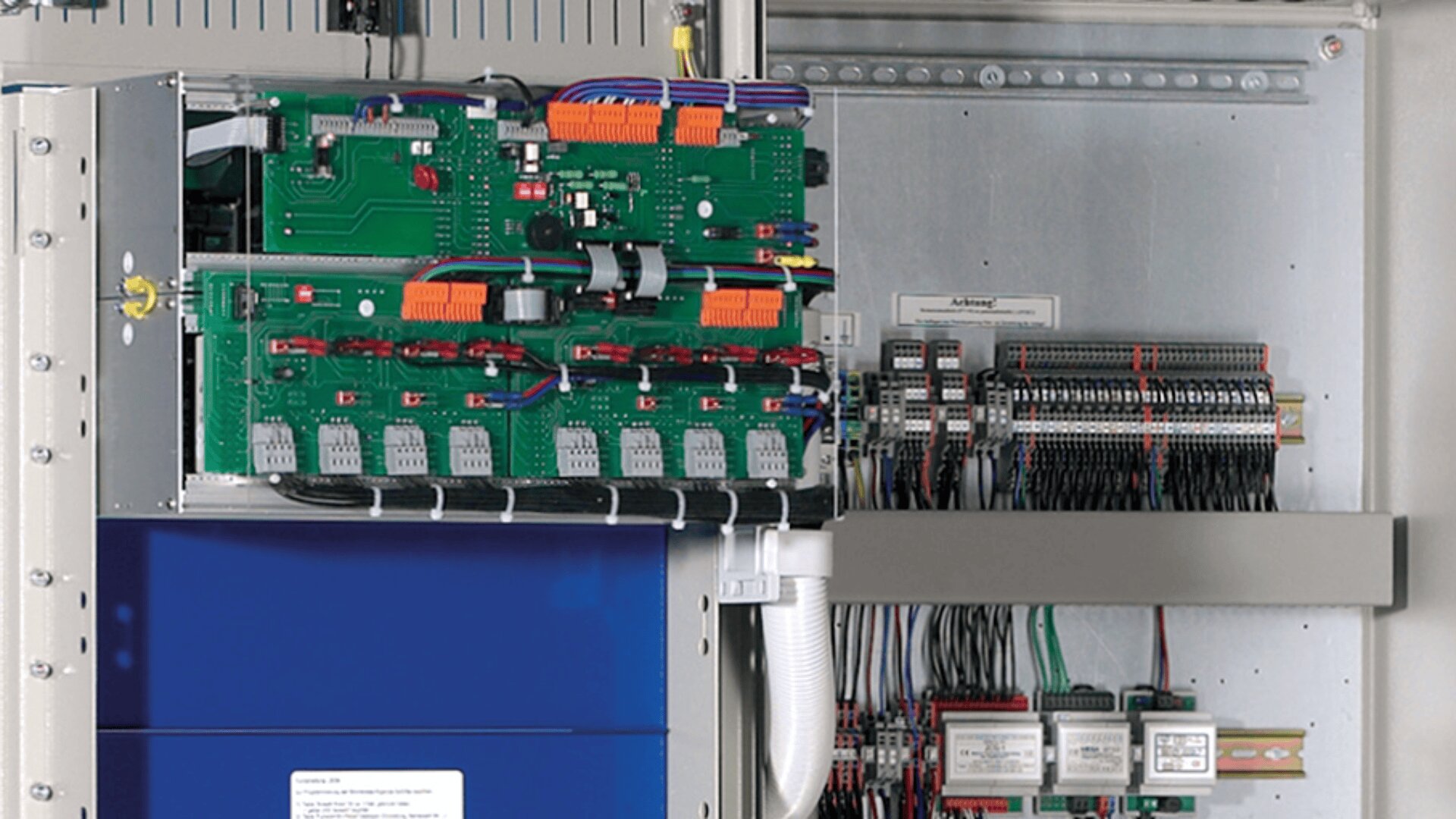 Eine geöffnete elektrische Schalttafel mit grünen Leiterplatten mit Drähten, Steckverbindern und Komponenten sowie Klemmenleisten und Relais, die in einem Metallgehäuse geordnet sind.