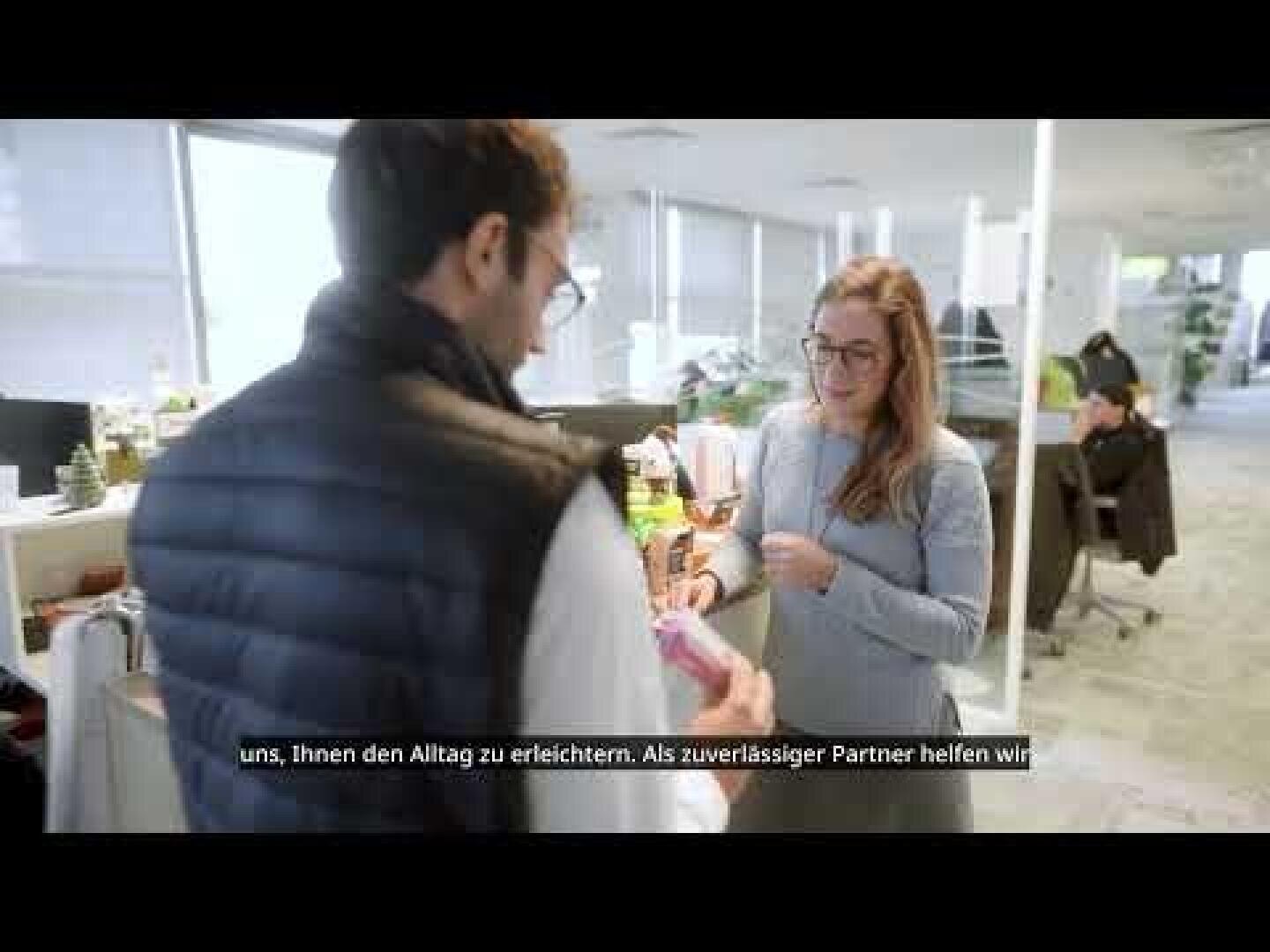 Ein Mann und eine Frau stehen in einem hellen, modernen Büro, unterhalten sich und halten Gegenstände in der Hand. Im Hintergrund arbeiten andere Personen an Schreibtischen. Am unteren Rand des Bildes erscheinen deutsche Untertitel.