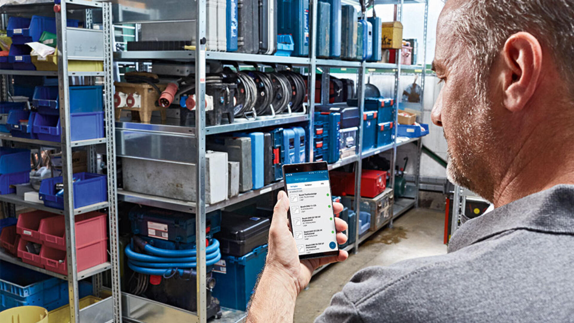 Un homme se tient dans un atelier ou une salle de stockage avec des étagères métalliques remplies d'outils et d'équipements. Il tient un smartphone et regarde une liste ou un inventaire sur l'écran.