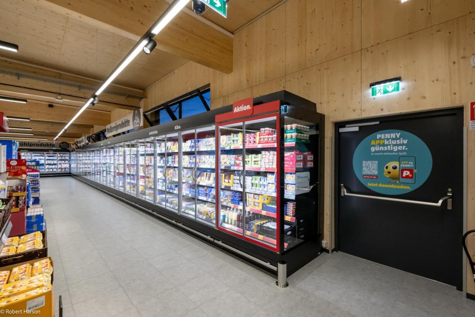 Une allée de supermarché très éclairée, avec des rayons réfrigérés regorgeant de produits variés et une porte noire sur laquelle est accrochée une affiche publicitaire ronde. Les murs et le plafond sont en bois.
