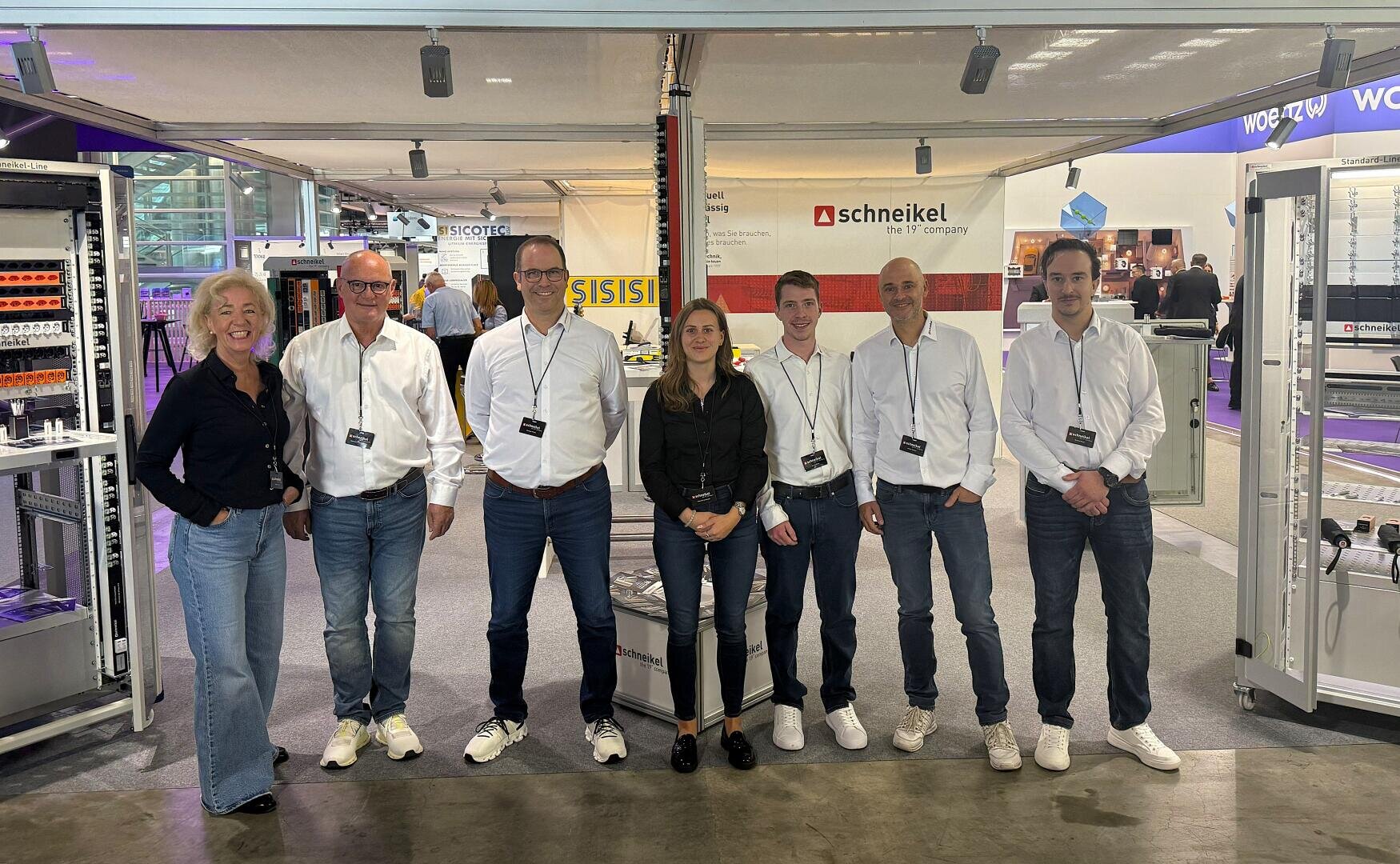 Sieben Personen – sechs Männer und eine Frau – stehen gemeinsam an einem Messestand, an dem Schalttafeln und elektrische Geräte ausgestellt sind. Alle tragen Namensschilder und sind leger oder im Business-Casual-Stil gekleidet; im Hintergrund sind Ausstellungsständer zu sehen.