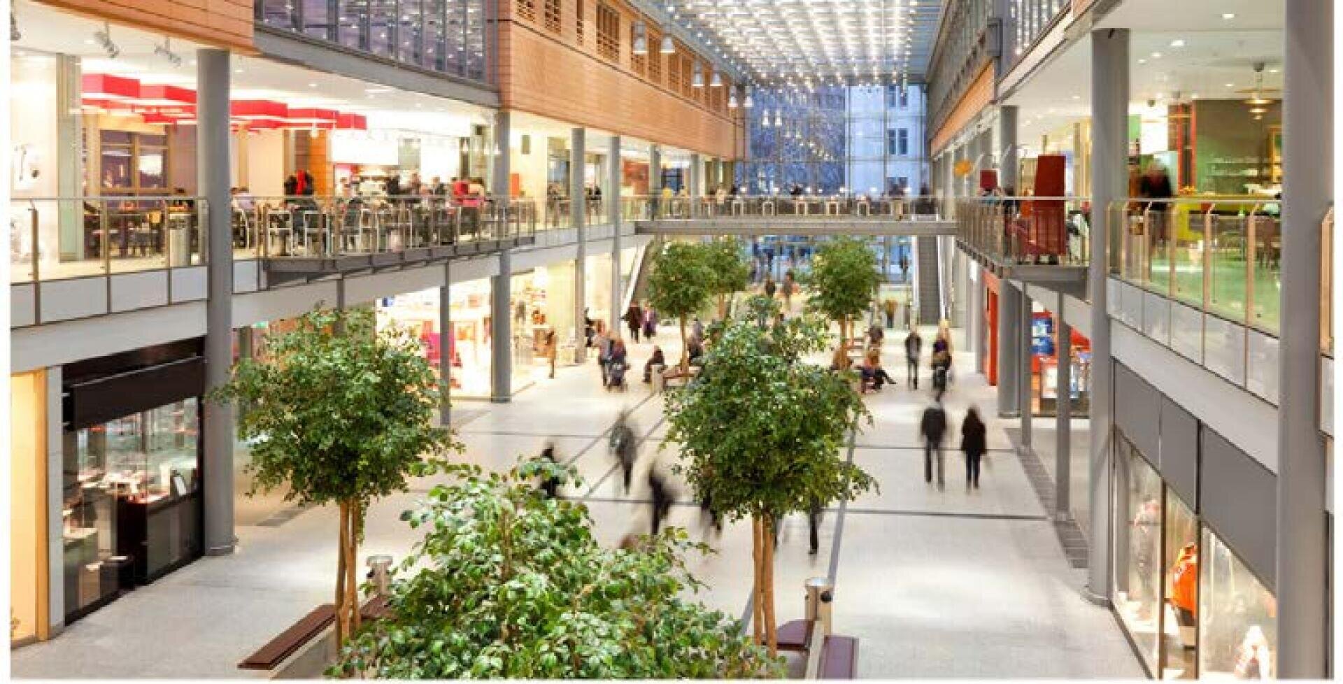Ein Einkaufszentrum mit zwei Etagen, Geschäften und einem Gastronomiebereich. Im Erdgeschoss schlendern die Menschen unter einer Glasdecke, durch die Tageslicht fällt, zwischen grünen Topfpflanzen umher.