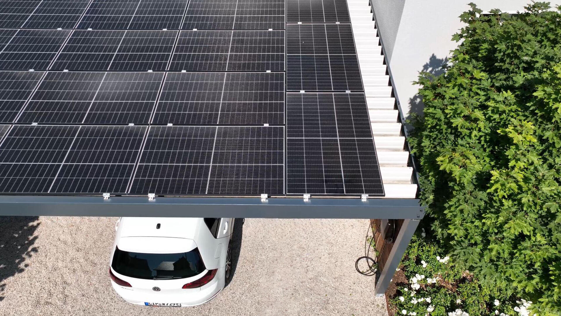 Une voiture blanche est garée sous un abri de voiture dont le toit est constitué de panneaux solaires. Le sol est recouvert de gravier et de la verdure avec des buissons entoure la zone sur le côté droit.