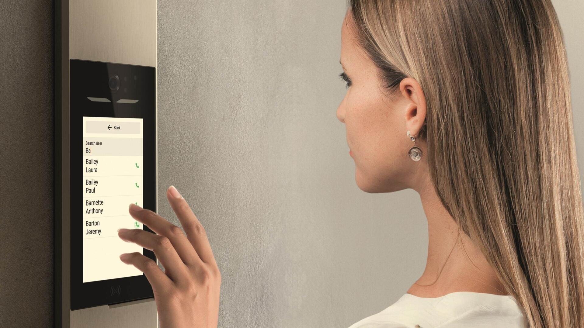 Eine Frau mit langen Haaren benutzt ein an der Wand montiertes Touchscreen-Gerät mit einer Liste von Namen und Telefonsymbolen, die suggerieren, dass sie jemanden anruft oder auswählt, den sie kontaktieren möchte. Das Gerät scheint eine intelligente Gegensprechanlage oder ein Sicherheitssystem zu sein.