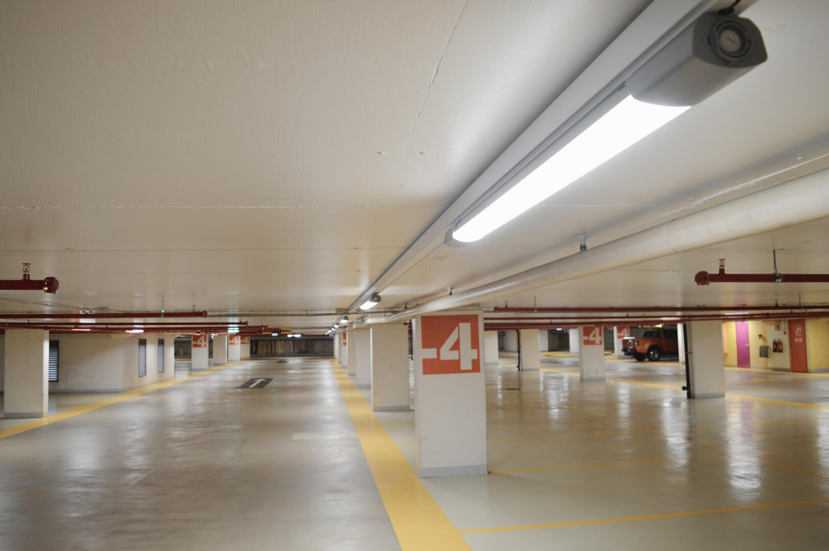 Ein leerer, gut beleuchteter Innenparkplatz mit weißen Wänden, gelben Linien und Säulen, die mit "-4" gekennzeichnet sind. Im Hintergrund sind einige geparkte Autos zu sehen.