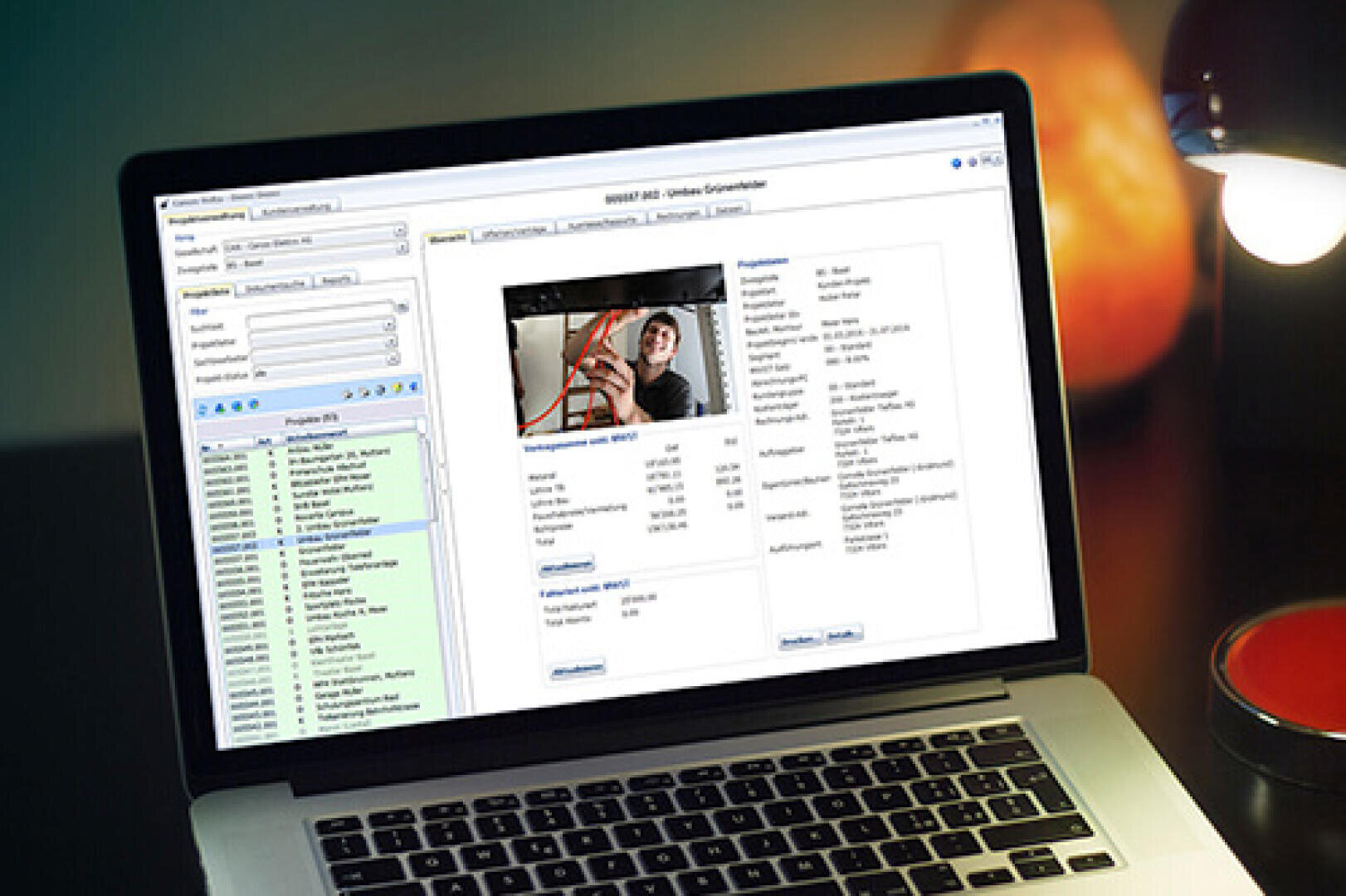 L'écran d'un ordinateur portable affiche une interface de base de données comportant une liste de noms, la photo d'une personne et des champs contenant des informations personnelles détaillées. Une lampe de bureau et un cahier rouge sont visibles à côté de l'ordinateur portable.