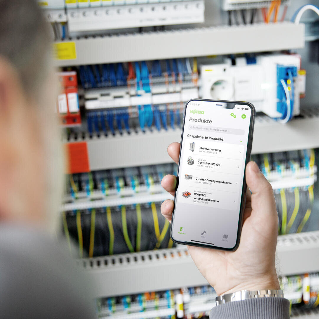 Une personne tient un smartphone, sur lequel s'affiche une application présentant une liste de produits, devant une armoire électrique ouverte contenant divers câbles et composants.