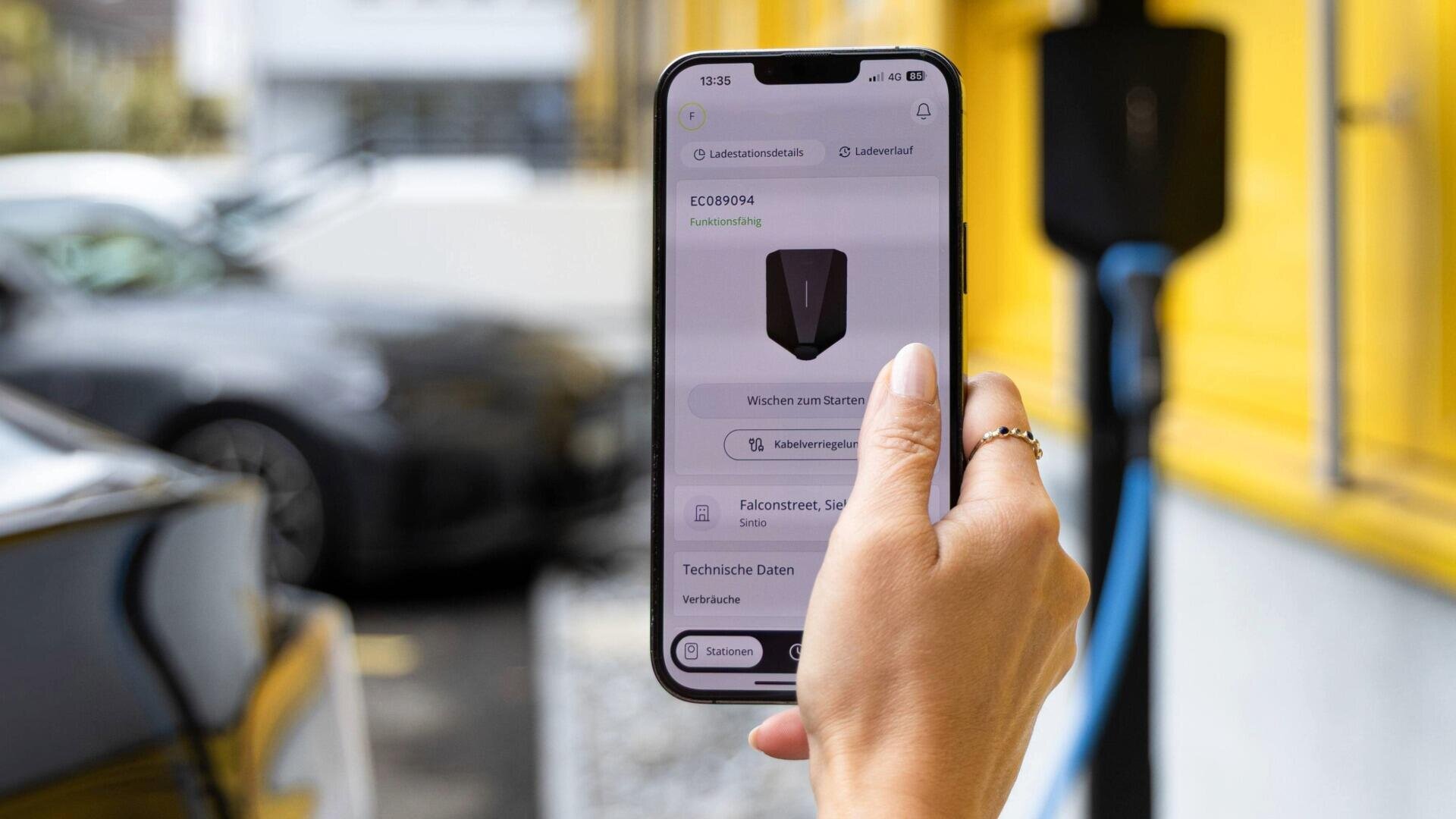 Eine Hand hält ein Smartphone, auf dem eine Lade-App für Elektrofahrzeuge geöffnet ist; das Smartphone befindet sich in der Nähe eines schwarzen Autos, das an einer Ladestation vor einer gelben Wand angeschlossen ist.