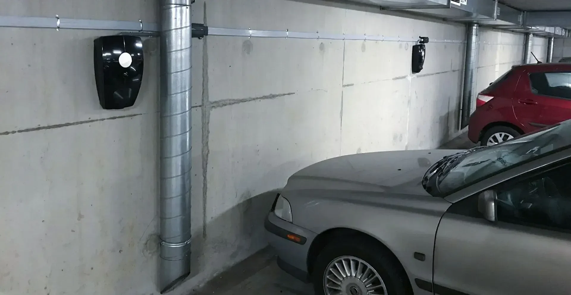 Une voiture argentée est garée dans un garage couvert, près d'un mur en béton sur lequel sont fixés deux appareils noirs. Une voiture rouge est garée à l'arrière-plan.