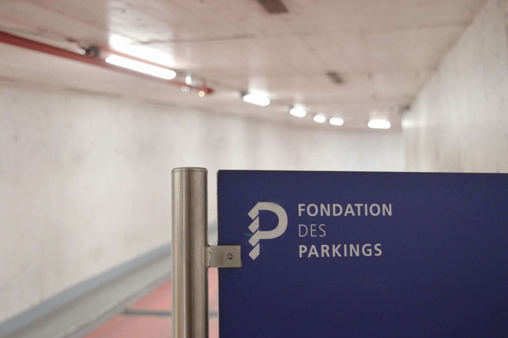 Ein blaues Schild mit der Aufschrift Fondation des Parkings in französischer Sprache ist an einem Metallpfosten im Inneren einer Tiefgarage oder eines Tunnels mit weißen, gut beleuchteten Wänden angebracht.
