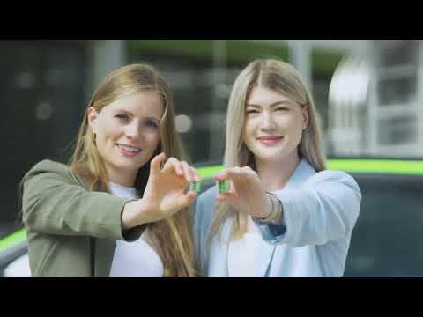 Zwei lächelnde Frauen stehen nebeneinander und halten jeweils einen grünen Autoschlüssel in Richtung Kamera, im Hintergrund ist ein grünes Auto zu sehen.