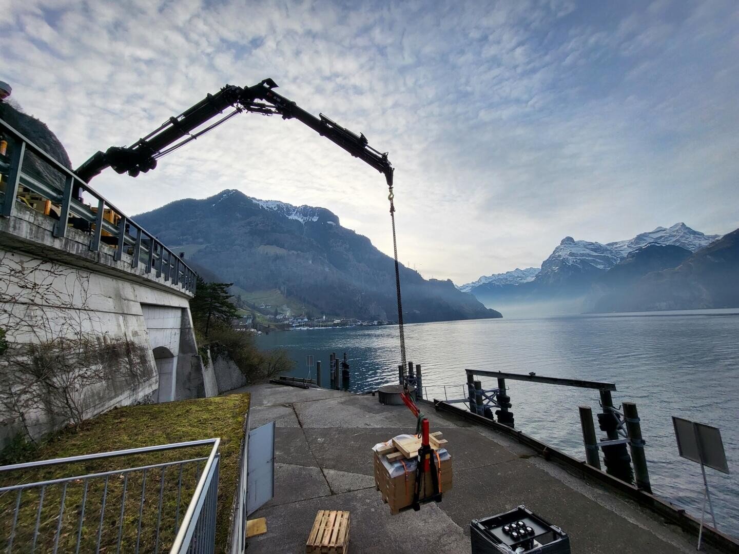 Sous un ciel nuageux, une grue soulève une charge près d'un lac entouré de montagnes ; des caisses et des palettes sont empilées sur le quai en béton.