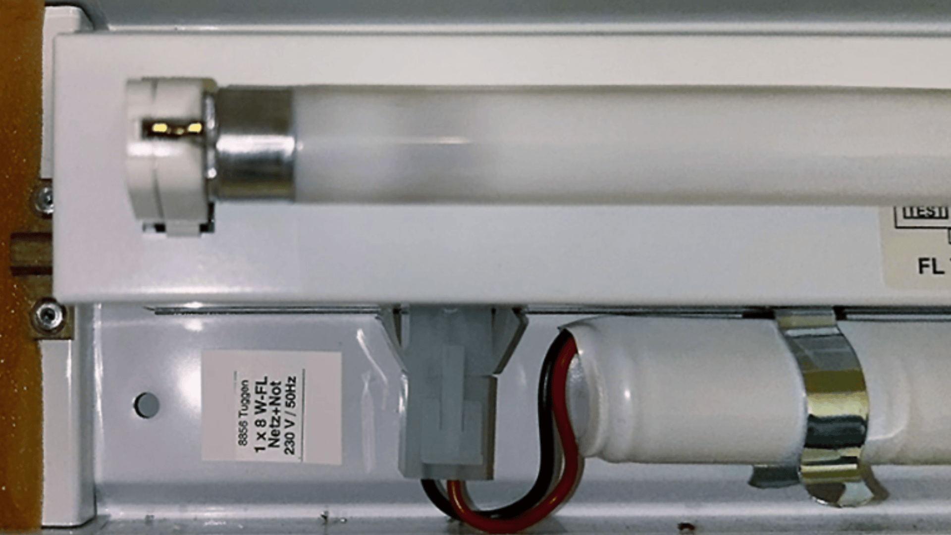 Nahaufnahme einer Leuchtstoffröhrenleuchte mit Lampe, elektrischer Verkabelung und einem weißen Etikett mit technischen Daten auf dem Metallsockel.
