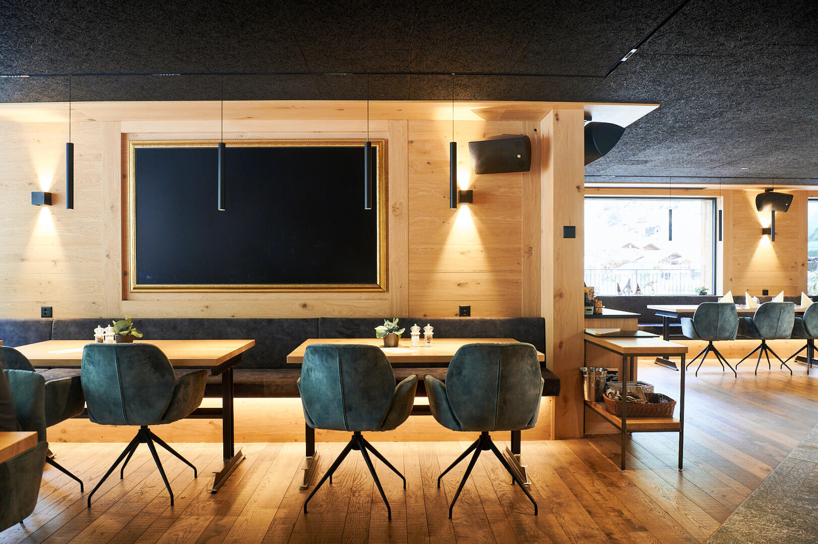 Modernes Restaurantinterieur mit Holzböden und -wänden, blaugrün gepolsterten Stühlen und Holztischen. An der Wand hängt eine große, leere Kreidetafel mit schwarzem Rahmen, und durch die Fenster im Hintergrund fällt Tageslicht herein.