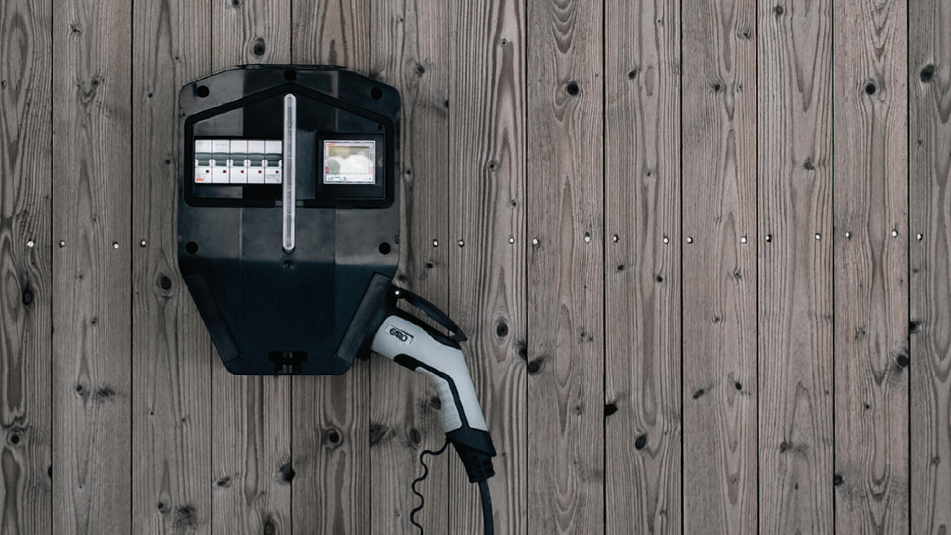 Eine schwarze Ladestation für Elektrofahrzeuge ist an einer Holzwand angebracht, an der ein Ladekabel und ein Stecker befestigt sind. Der Hintergrund besteht aus vertikalen Holzbrettern.