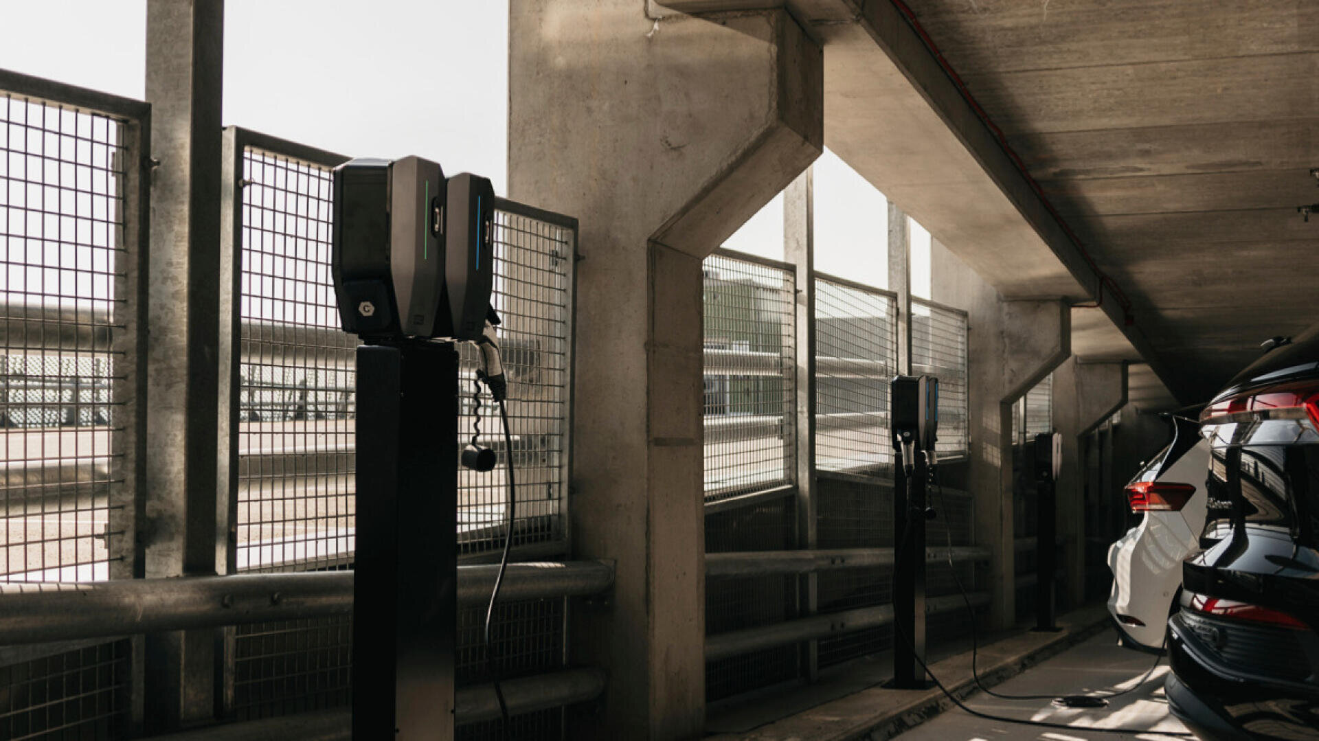 In einem Parkhaus aus Beton sind Ladestationen für Elektrofahrzeuge installiert, an deren Wänden mehrere Autos geparkt und an die Ladegeräte angeschlossen sind. Das Sonnenlicht fällt durch die Metallgitter auf der linken Seite.