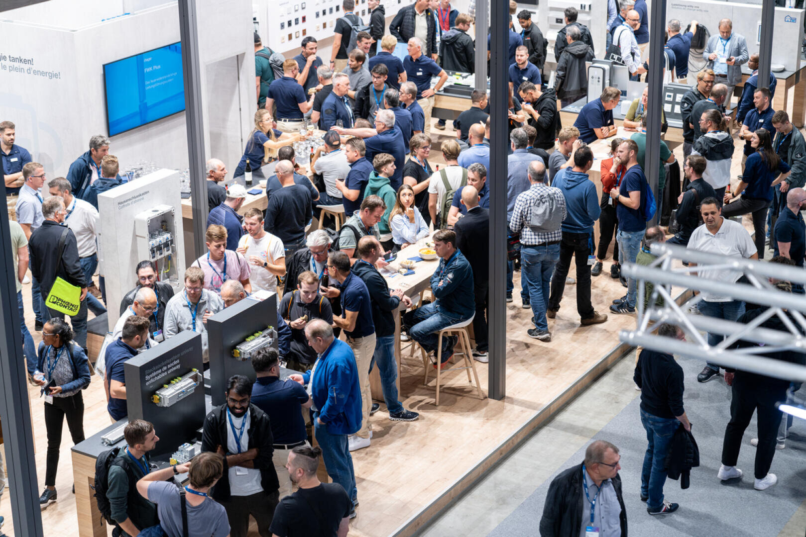 Eine große Gruppe von Menschen versammelt sich in einer belebten Ausstellungshalle, führt Gespräche und begutachtet Displays und elektronische Geräte an verschiedenen Ständen. Einige sitzen an Tischen, während andere stehen und durch den Raum gehen.