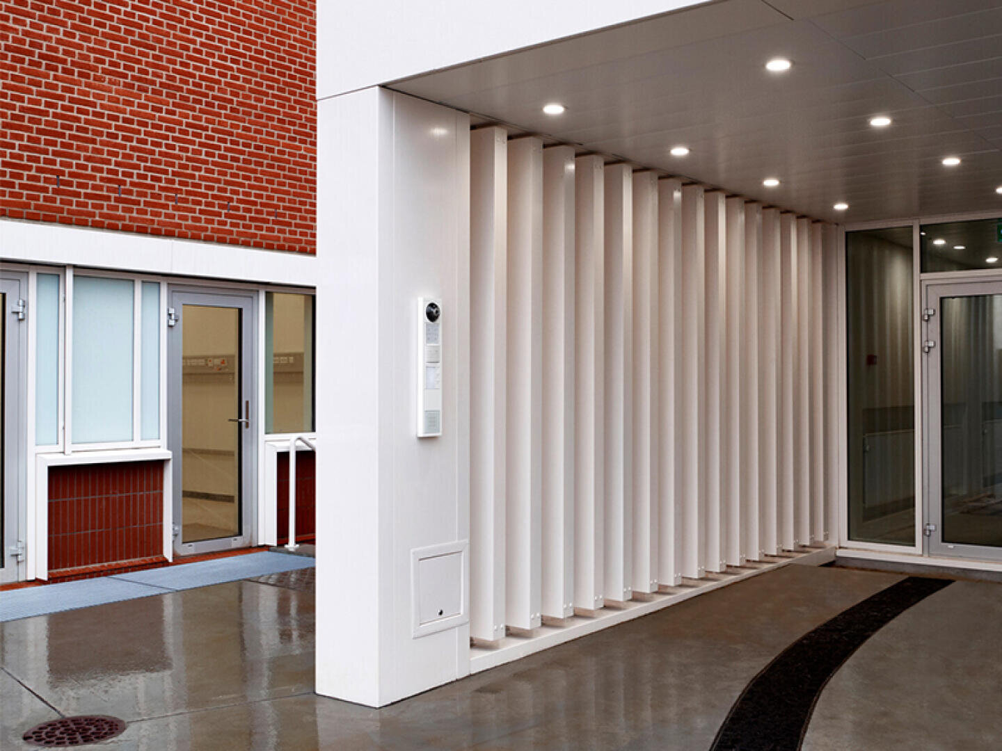 Entrée moderne d'un immeuble avec un passage couvert, un revêtement vertical blanc, des portes vitrées, un mur en briques rouges et un sol en béton encore humide, signe d'une pluie récente. Les plafonniers sont allumés et créent une atmosphère claire et élégante.