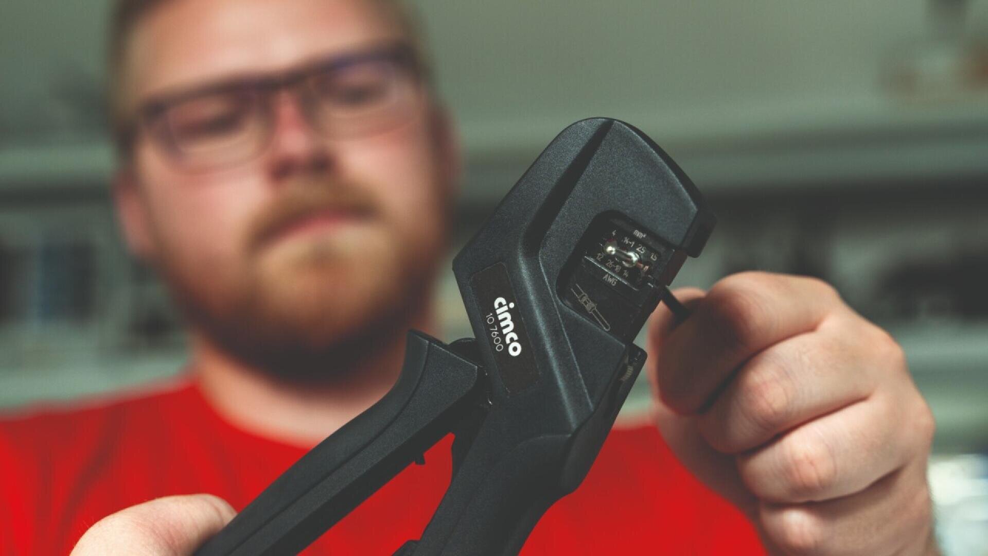 Une personne portant une barbe et des lunettes tient et utilise une pince à dénuder CIMCO noire, en se concentrant sur le dénudage d'un fil, avec un arrière-plan flou.