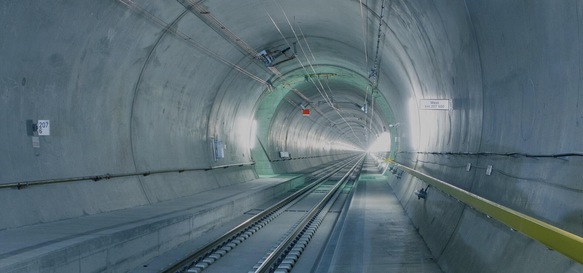 Ein langer, menschenleerer Eisenbahntunnel, durch dessen Mitte Stahlschienen verlaufen, der von künstlichem Licht beleuchtet wird und von grauen Betonwänden gesäumt ist. Auf der rechten Seite sind Schilder zu sehen, und der Tunnel verschwindet allmählich im grellen Licht in der Ferne.