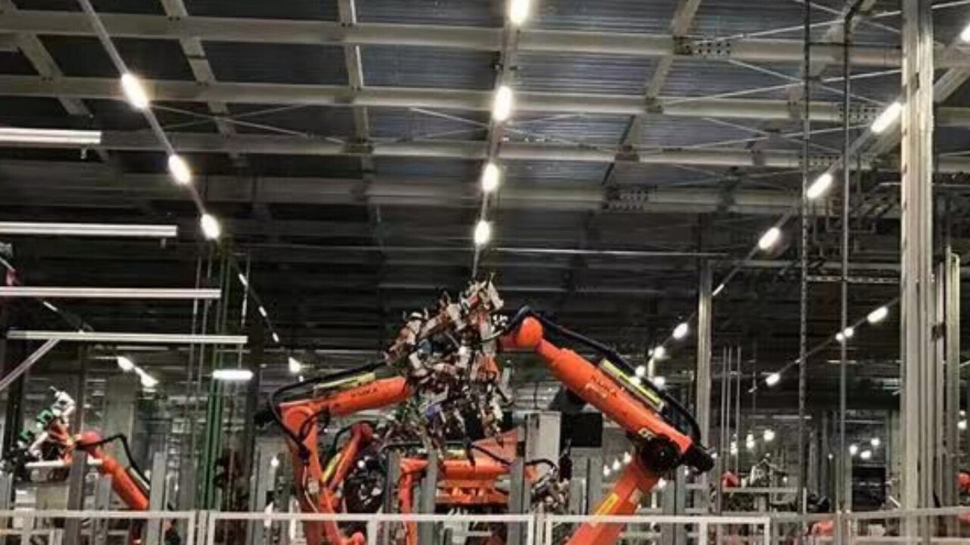Des bras robotisés orange travaillent ensemble dans une usine industrielle avec un éclairage zénithal, des structures métalliques et un haut plafond visible à l'arrière-plan.