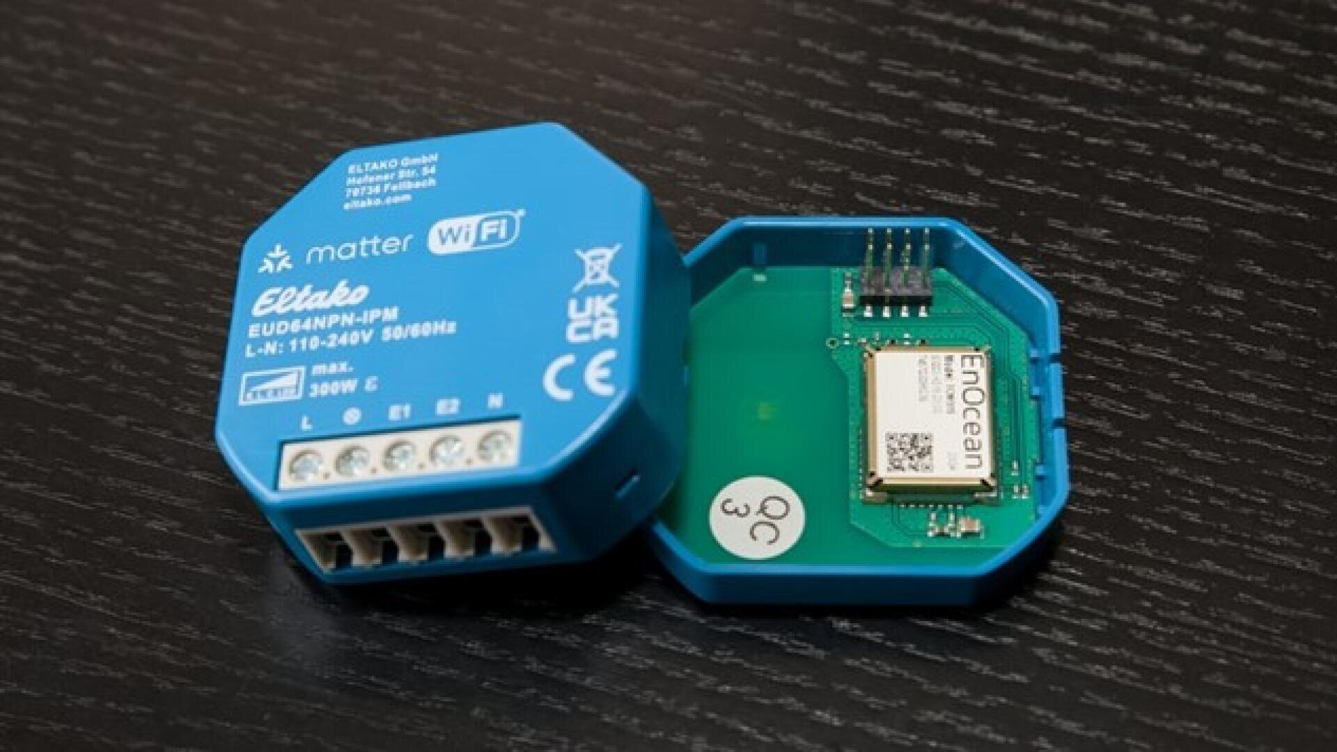 Ein blaues Eltako-Smart-Home-Modul, dessen eine Seite offen ist und das den Blick auf die interne Elektronik sowie einen beschrifteten EnOcean-Chip freigibt, auf einer dunklen Holzoberfläche platziert.