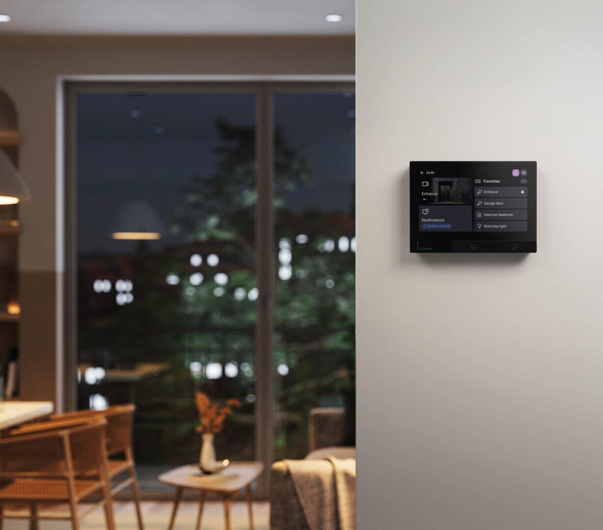 Un panneau de contrôle smart home est installé sur un mur blanc dans un salon moderne, avec une table à manger et des chaises au premier plan et de grandes portes vitrées montrant une vue extérieure nocturne.