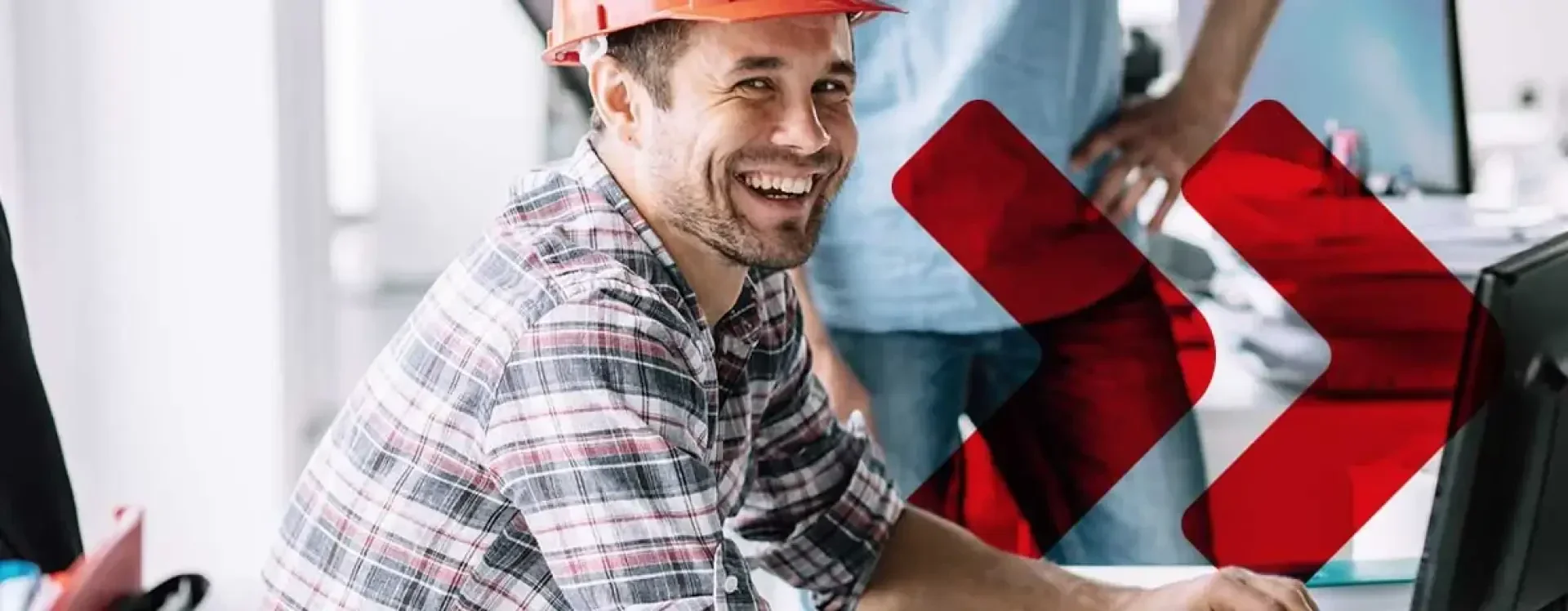 Un homme souriant, vêtu d'une chemise à carreaux et d'un casque de chantier orange, travaille devant un ordinateur dans un bureau ; deux flèches rouges sont superposées à l'image et une autre personne se tient debout à l'arrière-plan.