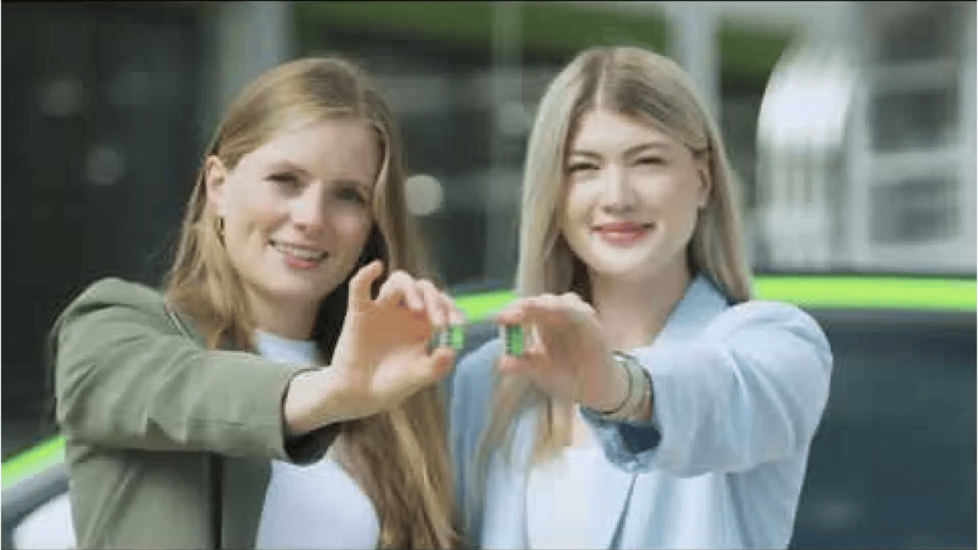 Deux femmes souriantes, debout à l'extérieur, tiennent ensemble un objet vert devant elles. Une voiture avec une bande verte est visible à l'arrière-plan.