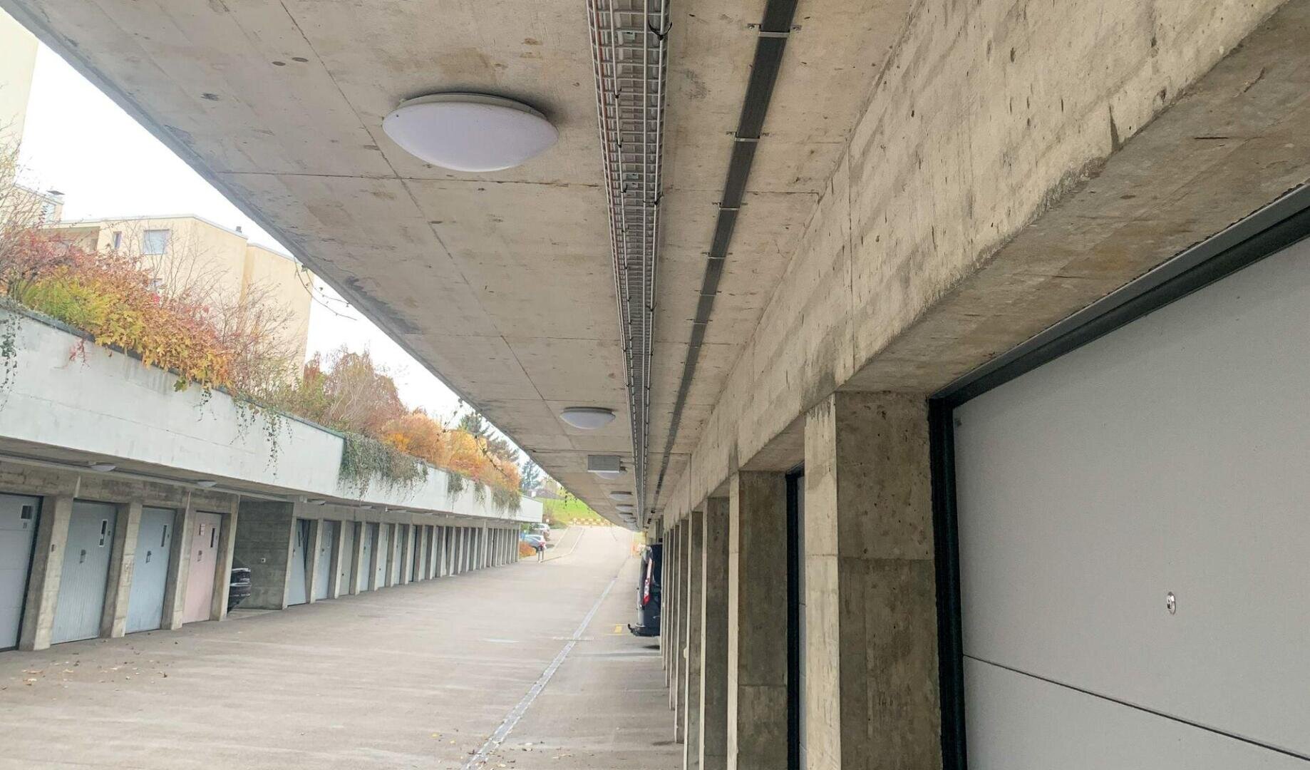 Eine Reihe grauer Garagentore unter einem Betonvordach, mit an der Decke befestigten Außenleuchten und sichtbarer Bepflanzung auf dem Dach, links.