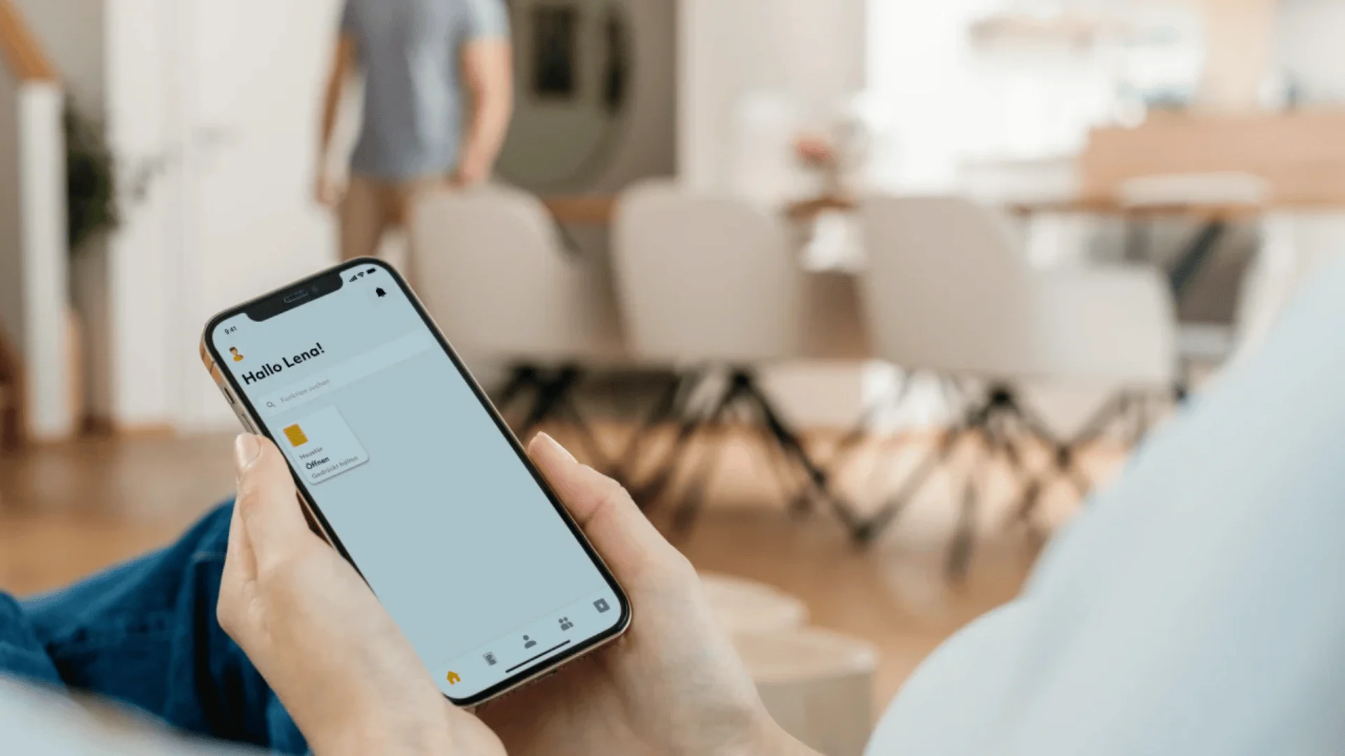 Person, die ein Smartphone hält, auf dem ein Faltdisplay mit der Aufschrift "Hallo, Lena!" angezeigt wird, in einem hellen, modernen Wohnzimmer mit einer unscharfen Person, die im Hintergrund läuft.