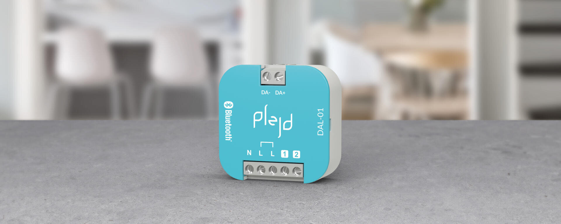 Ein kleines blau-weißes elektronisches Gerät mit der Aufschrift „plejd“ und „Bluetooth“ steht auf einer grauen Fläche vor einem unscharfen Innenraumhintergrund, auf dem Stühle und Tische zu sehen sind.