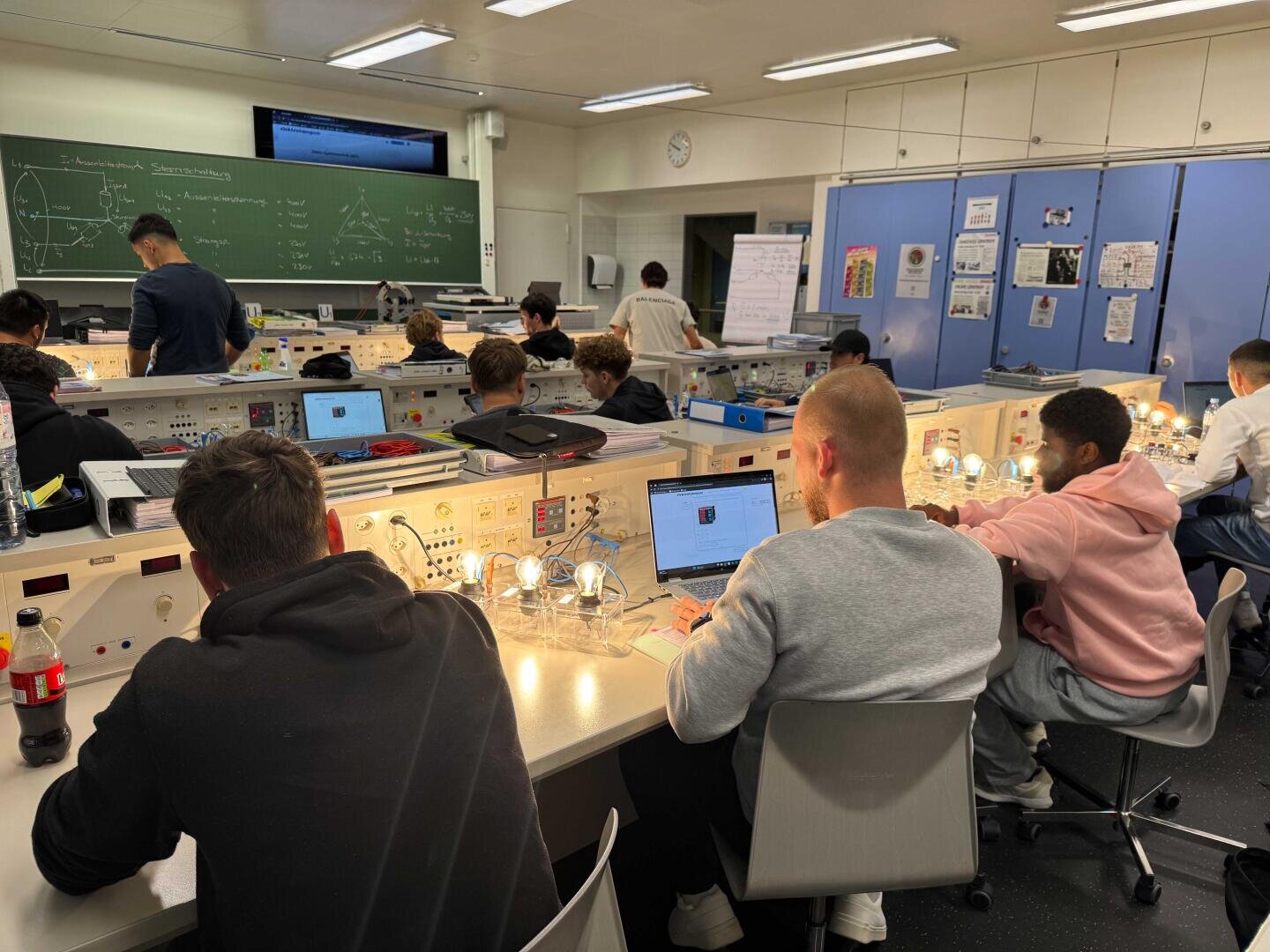 Des élèves travaillent sur des postes de laboratoire individuels avec des ordinateurs portables et des appareils éclairés dans une salle de classe lumineuse. Un enseignant se tient devant un tableau couvert de diagrammes et de notes. Des posters scientifiques et des articles techniques sont accrochés aux murs.