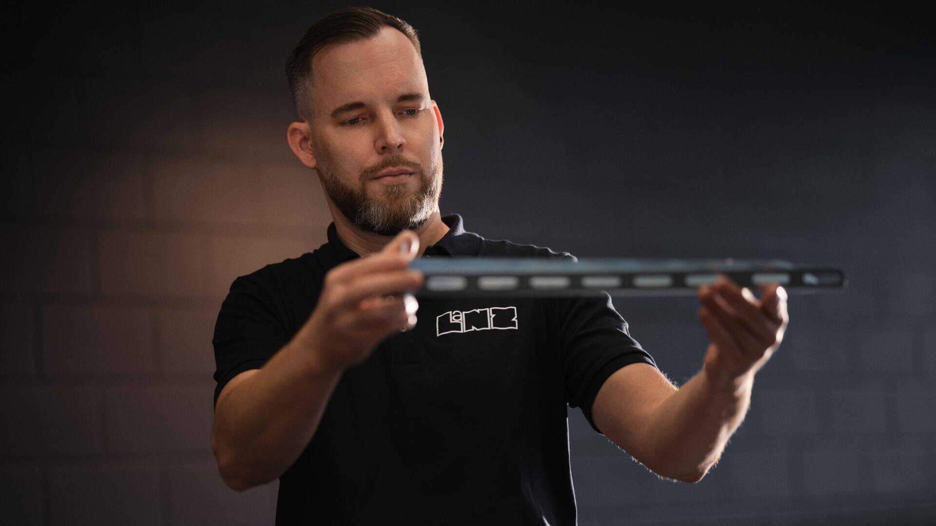 Ein bärtiger Mann in einem schwarzen Polohemd hält einen langen, dünnen Metallgegenstand waagerecht in der Hand und betrachtet ihn aufmerksam vor einem dunklen, unscharfen Hintergrund.