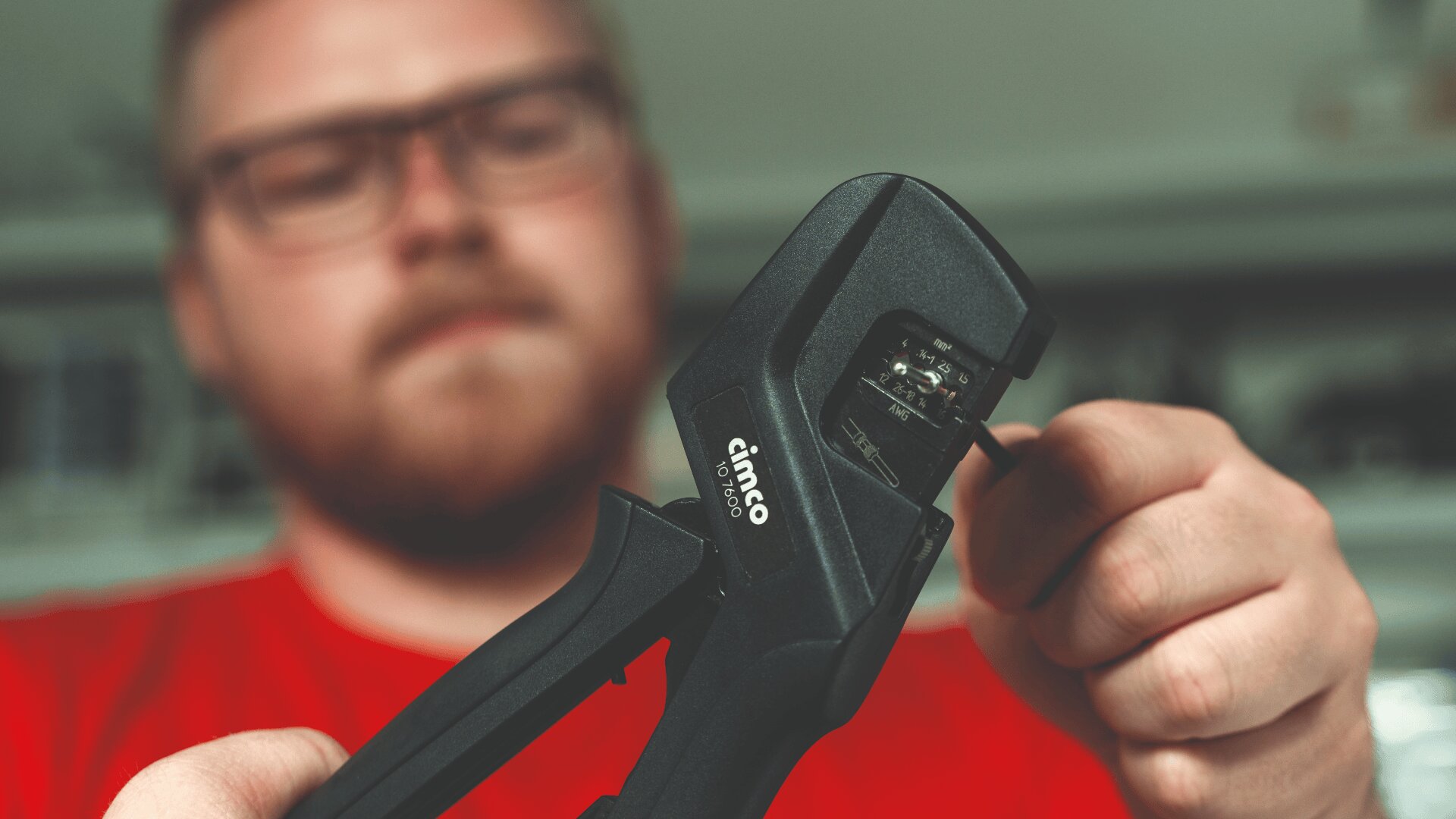 Eine Person mit Brille und rotem Hemd verwendet eine Cimco-Crimpzange und konzentriert sich auf die Einstellung oder Bedienung der Zange mit beiden Händen. Der Hintergrund ist unscharf, um die Aufmerksamkeit auf das Werkzeug und die Hände zu lenken.