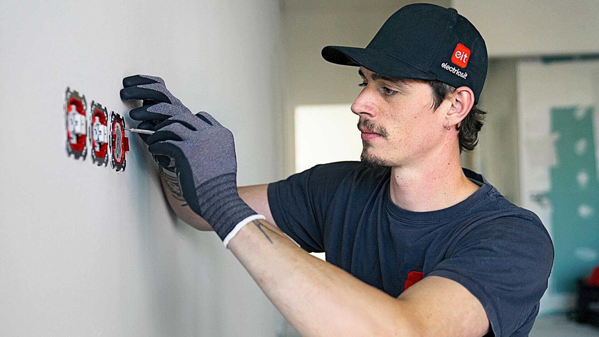 Eine Person, die Handschuhe und eine schwarze Mütze trägt, montiert oder justiert Steckdosen an einer weißen Wand in einem Raum, der gerade gebaut oder renoviert wird.