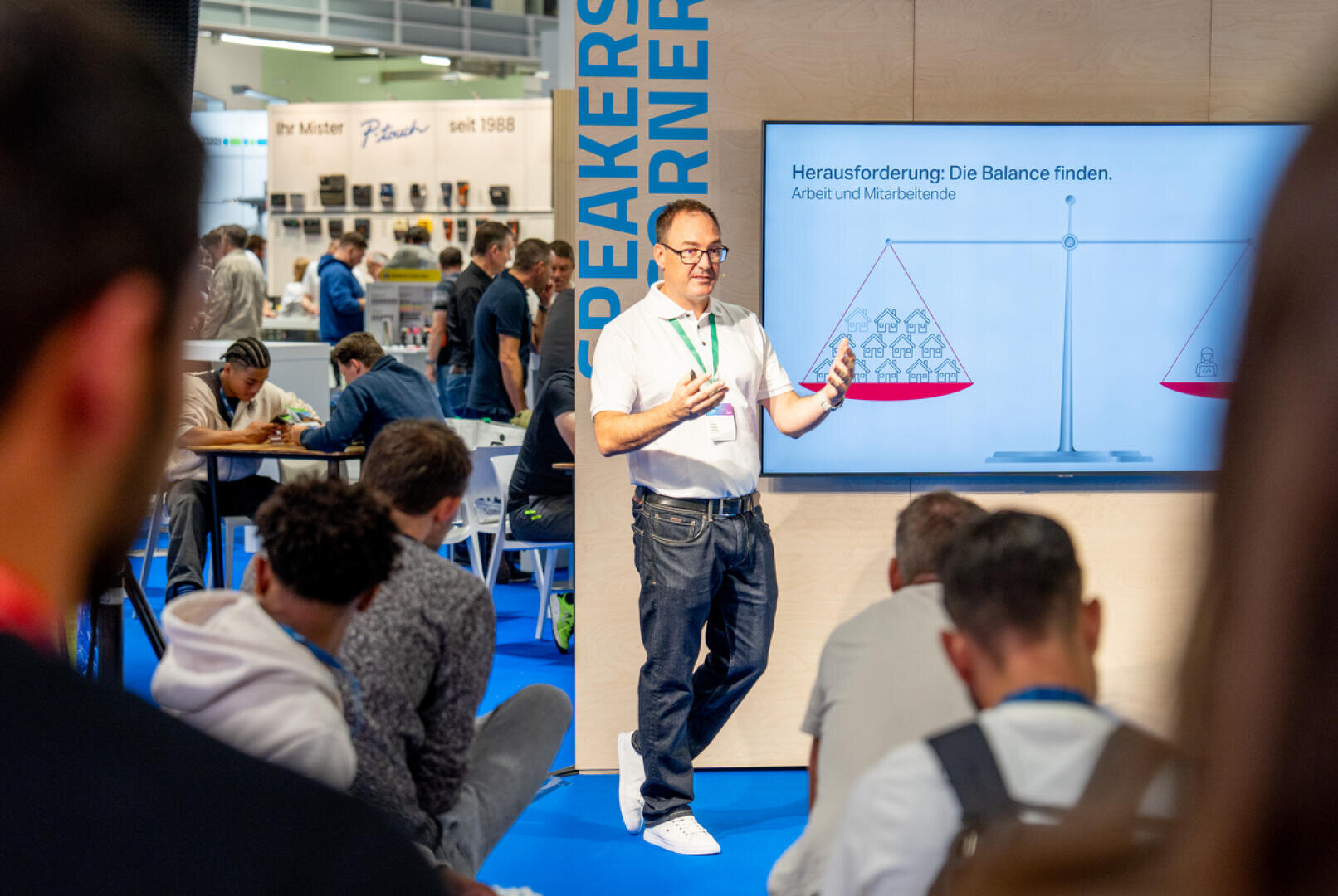 Ein Mann hält einen Vortrag vor einem Publikum auf einer Messe. Er steht vor einem Bildschirm, auf dem eine Waage zu sehen ist, die Arbeit und Material abwägt. Mehrere Personen sitzen und hören aufmerksam zu. Die Umgebung ist hell und geschäftig.