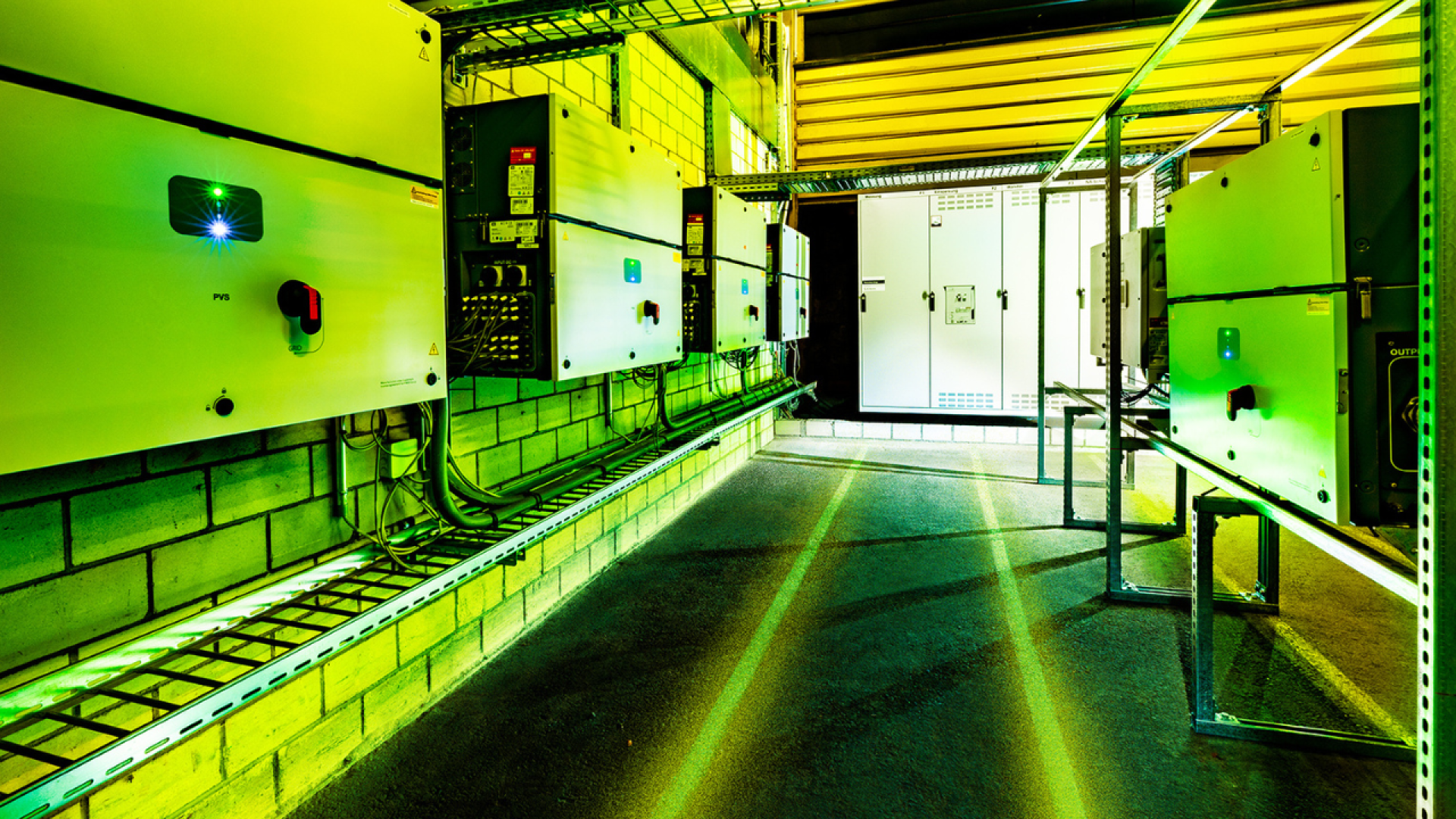 Hell erleuchteter Industrieraum mit grün getönter Beleuchtung, in dem elektrische Schalttafeln an Backsteinwänden und Kabeltrassen angebracht sind, wobei das Sonnenlicht durch eine teilweise geöffnete Tür im Hintergrund einfällt.