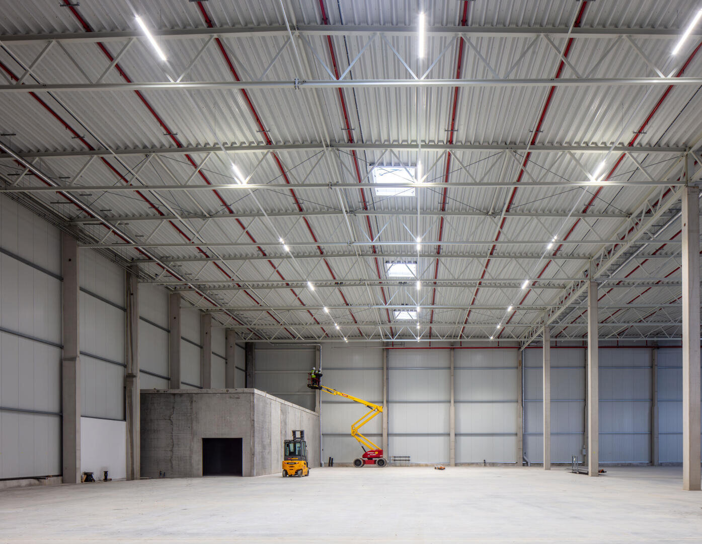 Un grand entrepôt industriel vide avec de hauts plafonds, des poutres métalliques et des lucarnes claires. Un ouvrier manipule une plate-forme élévatrice jaune près d'une petite structure en béton contre un mur.