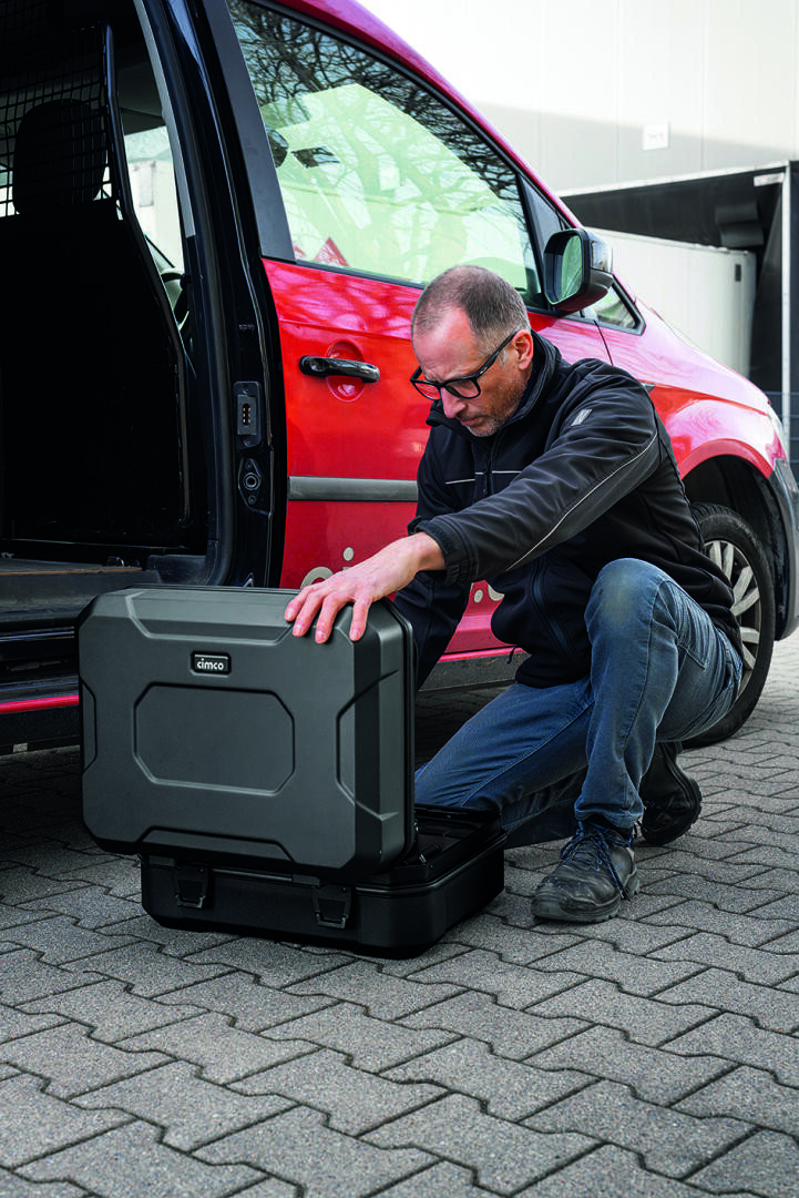 Un homme est agenouillé sur le trottoir, à côté d'une camionnette rouge, et ouvre une grande valise rigide noire posée par terre. La porte coulissante de la camionnette est ouverte, et l'homme semble entièrement concentré sur le contenu de la valise.