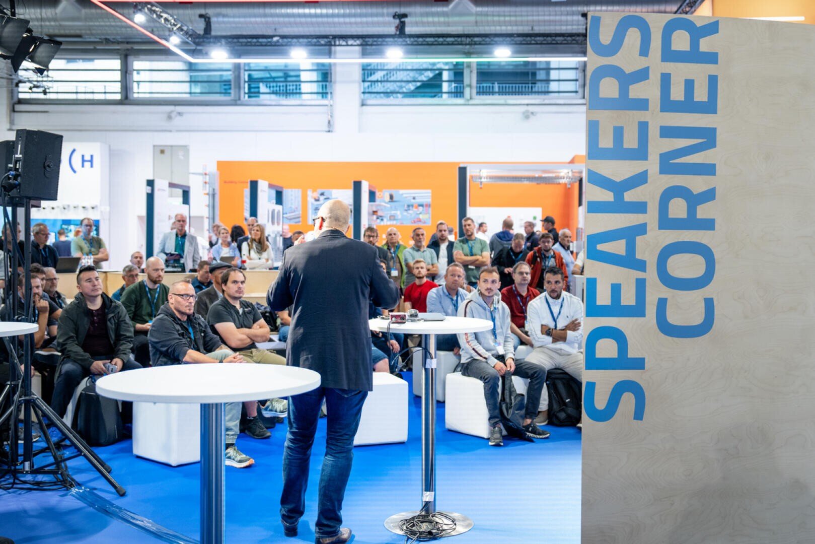 Ein Redner wendet sich bei einer Veranstaltung in einer Halle an ein aufmerksames Publikum. Er steht in der Nähe von Tischen und einer großen Wand, die mit SPEAKERS CORNER in blauer Schrift beschriftet ist. Das Publikum sitzt dem Redner gegenüber.