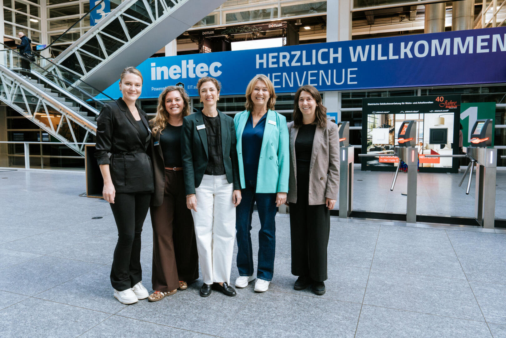 Cinq femmes souriantes se tiennent devant un panneau bleu indiquant « HERZLICH WILLKOMMEN / BIENVENUE » lors d'un événement en intérieur ; derrière elles, on aperçoit le logo ineltec et des expositions technologiques modernes.