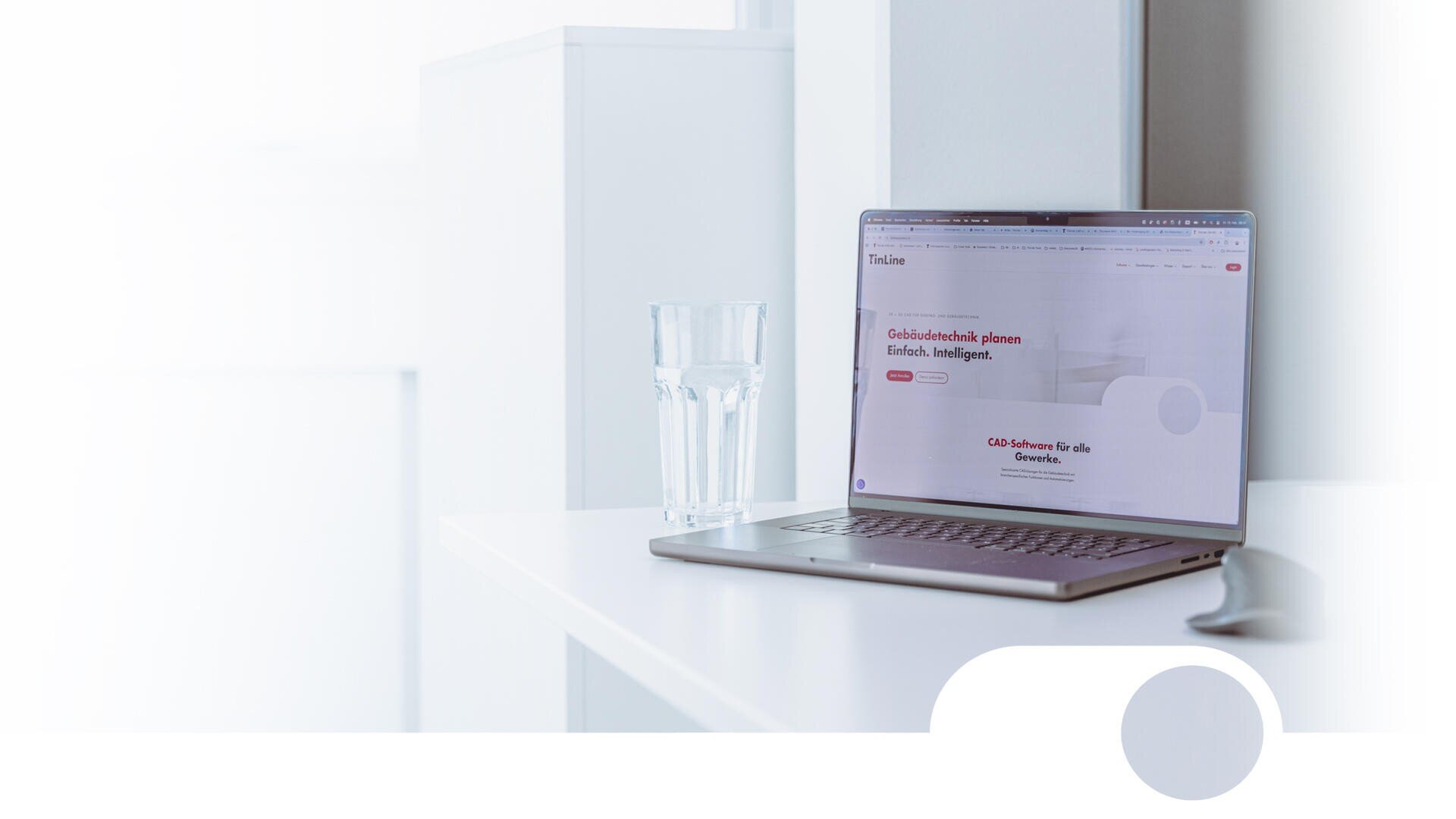 Un ordinateur portable est posé sur un bureau blanc, à côté d'un verre d'eau. L'écran de l'ordinateur affiche une page web où prédominent les couleurs blanche et rouge, avec du texte en allemand. L'arrière-plan et l'environnement sont lumineux et épurés.