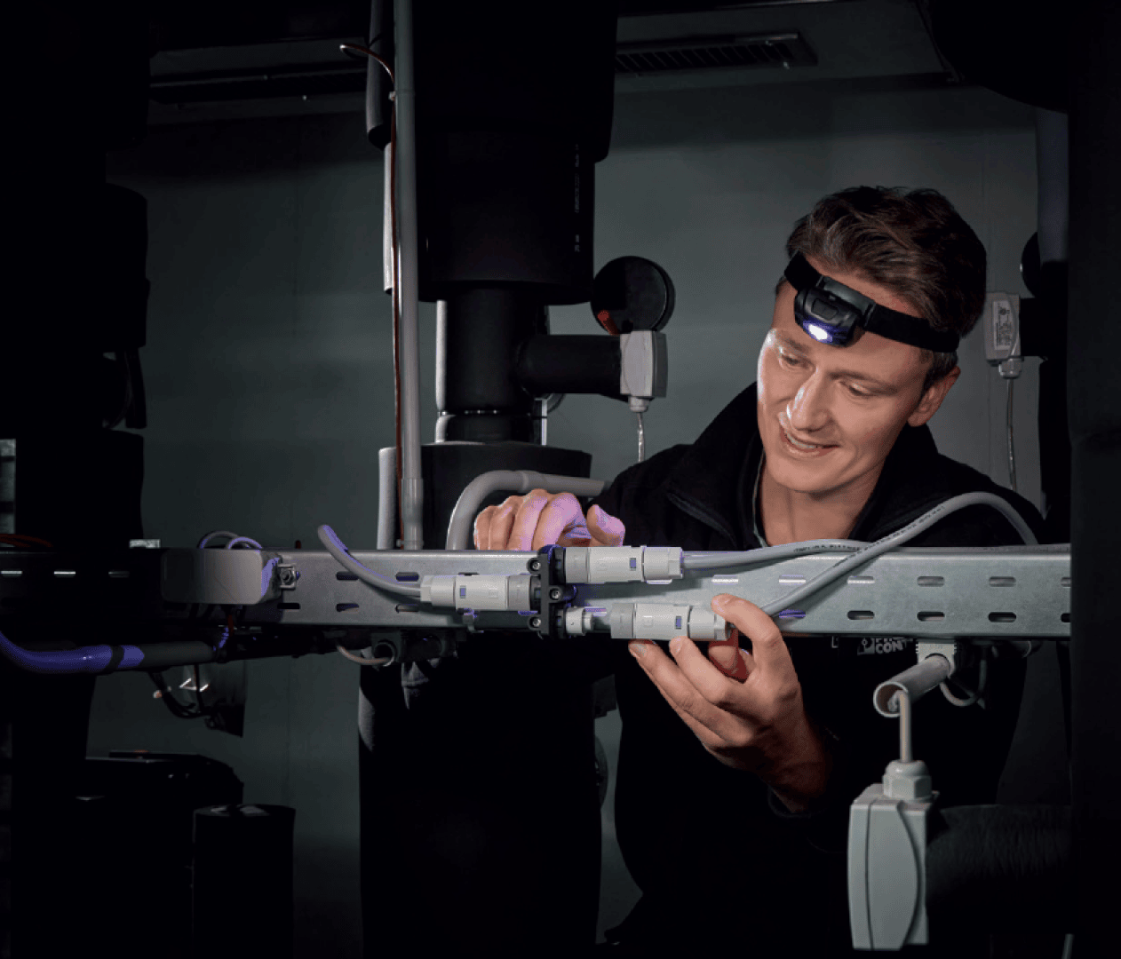 Eine Person mit einer Stirnlampe arbeitet in einem schwach beleuchteten Raum mit elektrischen Kabeln und Steckverbindern an einem Metallrahmen und konzentriert sich mit beiden Händen auf die Aufgabe.